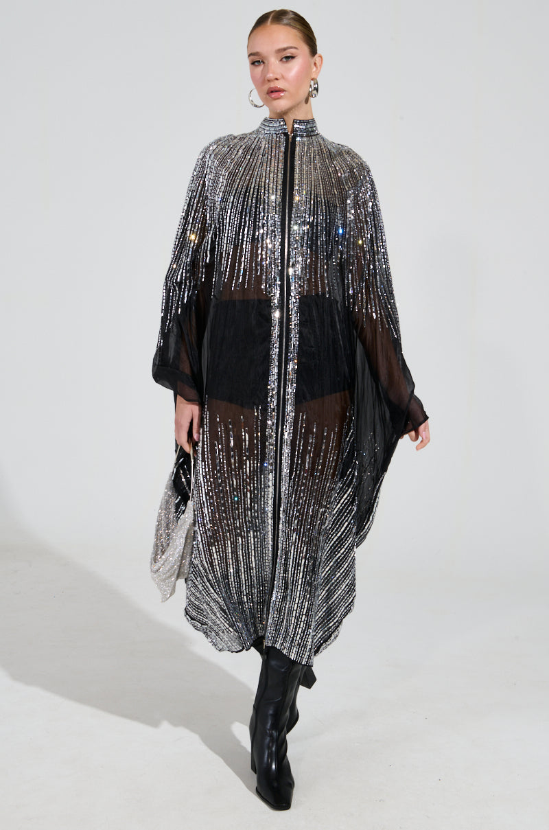 A model struts down the runway in the SPARKLE AND SHINE BLOUSE, featuring shimmering mirrored sequins cascading over a sheer nylon black dress. She accessorizes with a small matching clutch and steps confidently in tall black boots against a plain white backdrop.
