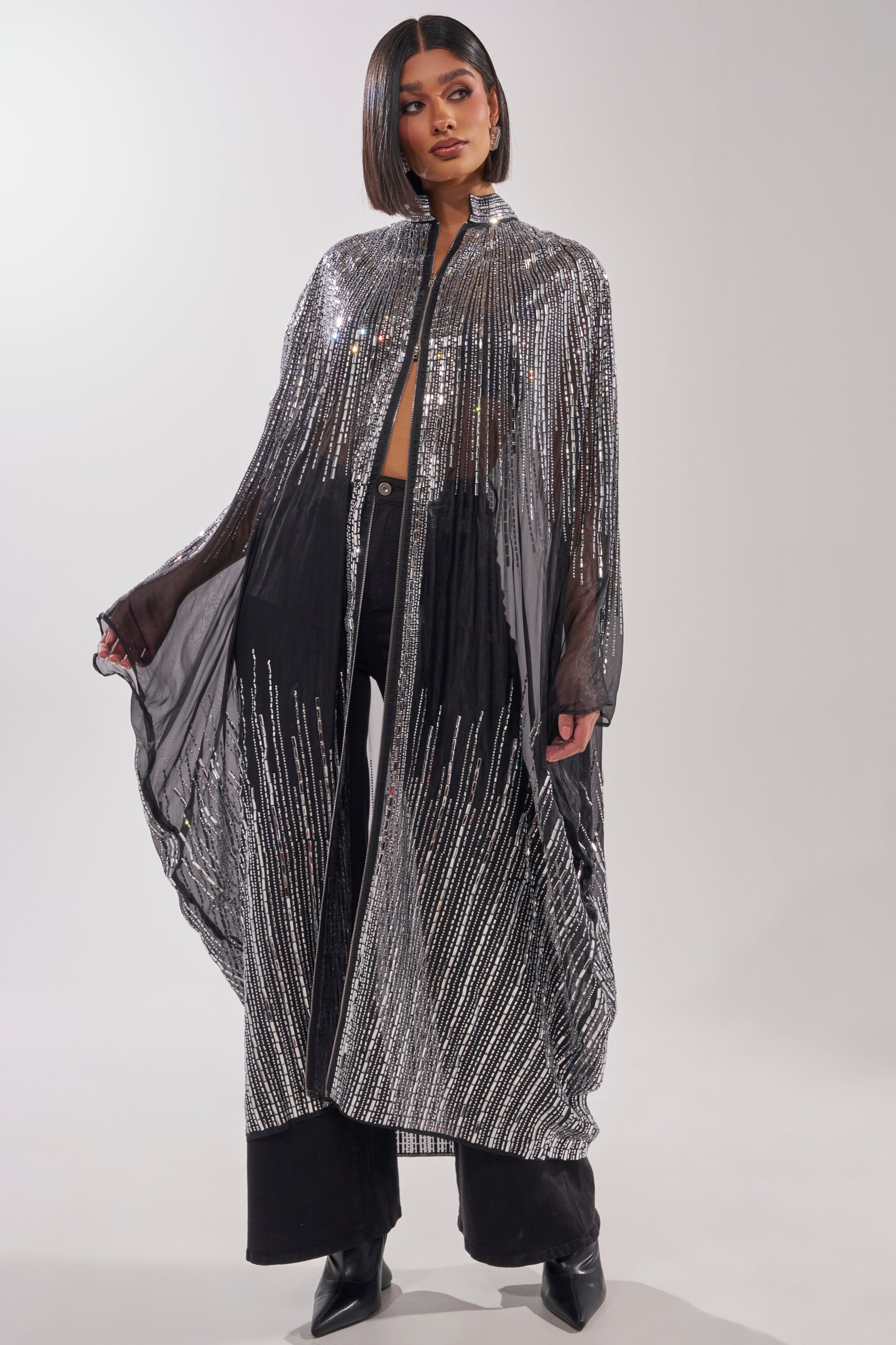 A woman stands in a studio wearing the SPARKLE AND SHINE BLOUSE under a long, sheer black cape with silver stripes, paired with black pants and boots. Her hair is styled in a sleek bob. 0