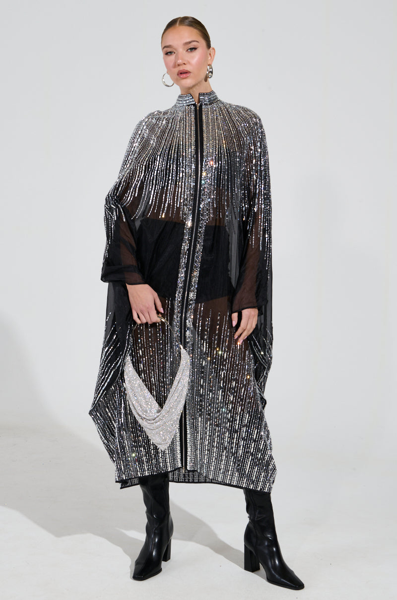 Dressed in the SPARKLE AND SHINE BLOUSE with its glistening metallic stripes and sheer nylon fabrication adorned with mirrored sequins, a person stands confidently. They enhance the outfit with knee-high black boots and large hoop earrings, their hair elegantly styled in a sleek updo.