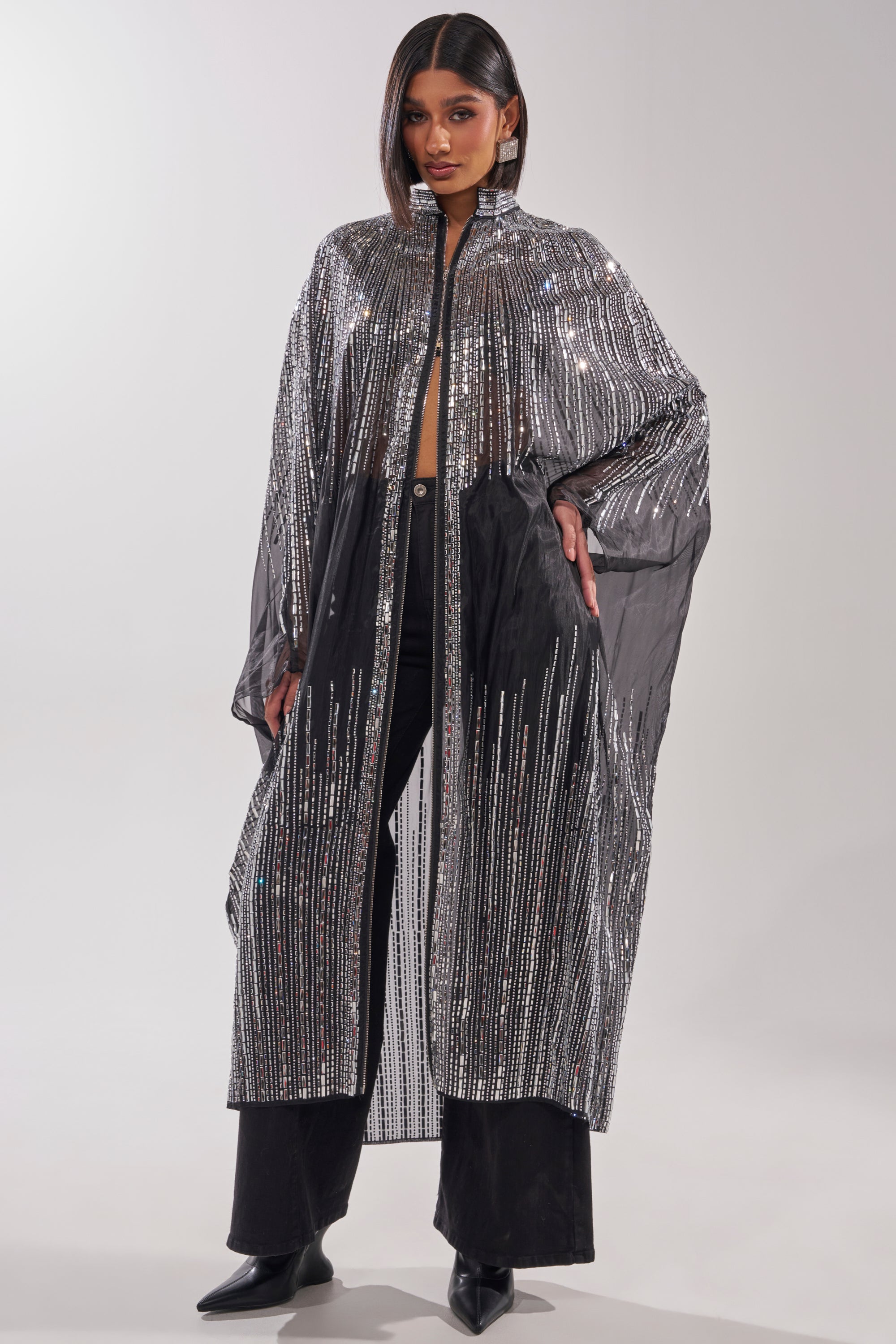 A woman with straight, shoulder-length hair poses against a light backdrop in the SPARKLE AND SHINE BLOUSE, a long, sheer black cover-up with vertical silver sequin stripes, styled over black pants and top with black heeled boots. 1