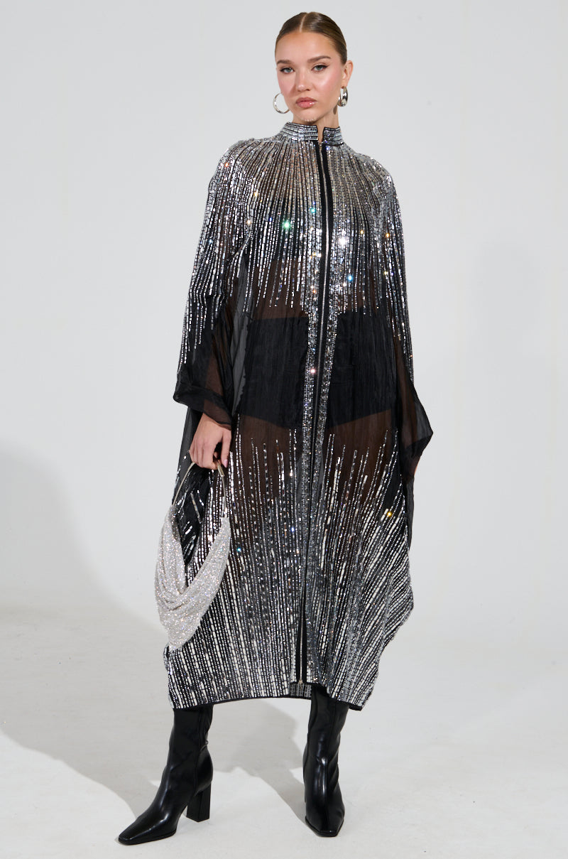Dressed in a dazzling SPARKLE AND SHINE BLOUSE over a sheer black dress crafted from transparent nylon, a person stands confidently. They're strikingly accessorized with knee-high black boots, a matching sequined bag, and hoop earrings against a plain backdrop.