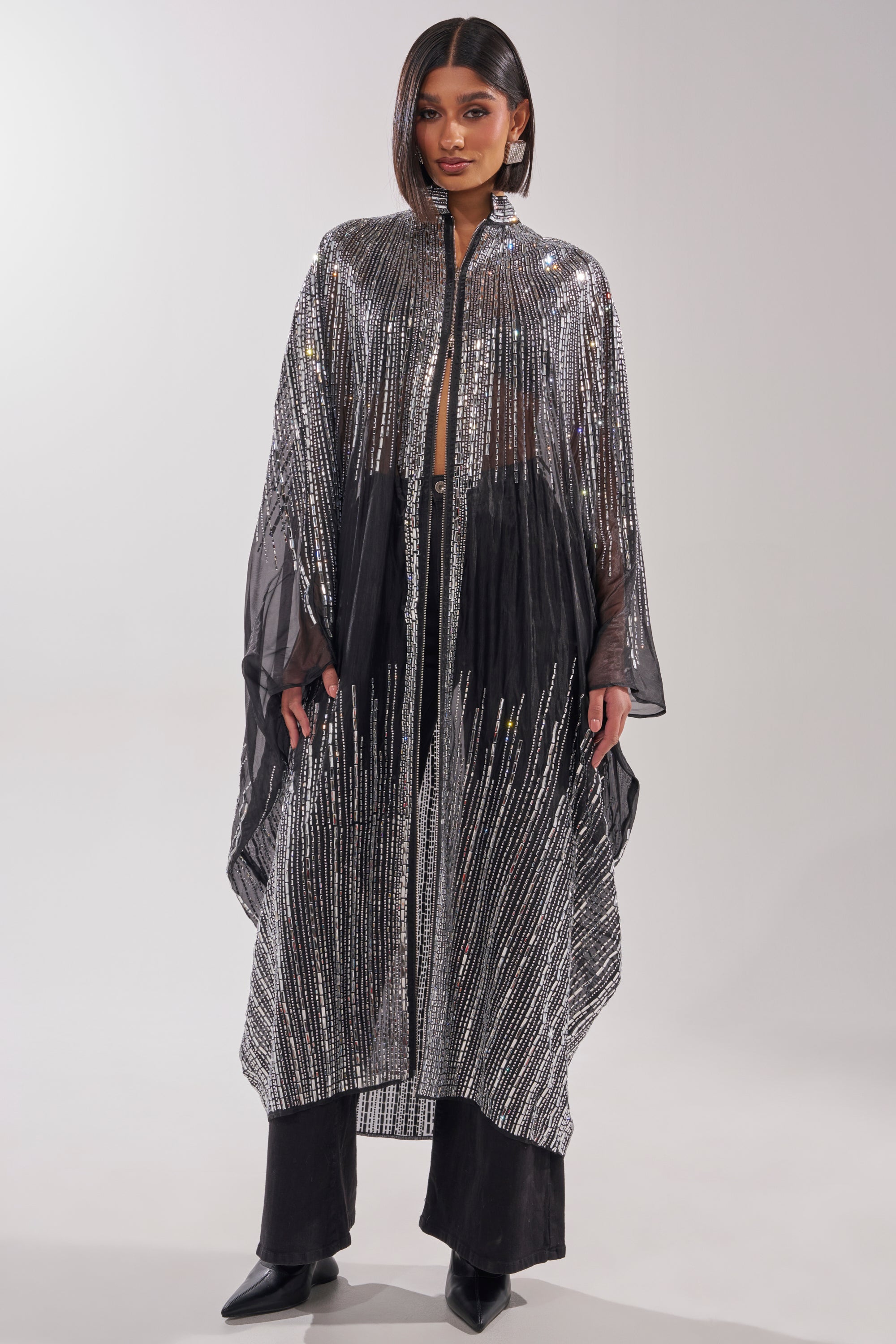 A person stands facing forward in a sheer, long black cape with silver stripes and sequins, layered over the SPARKLE AND SHINE BLOUSE, black pants, and pointed-toe shoes. The background is plain and light-colored. 2