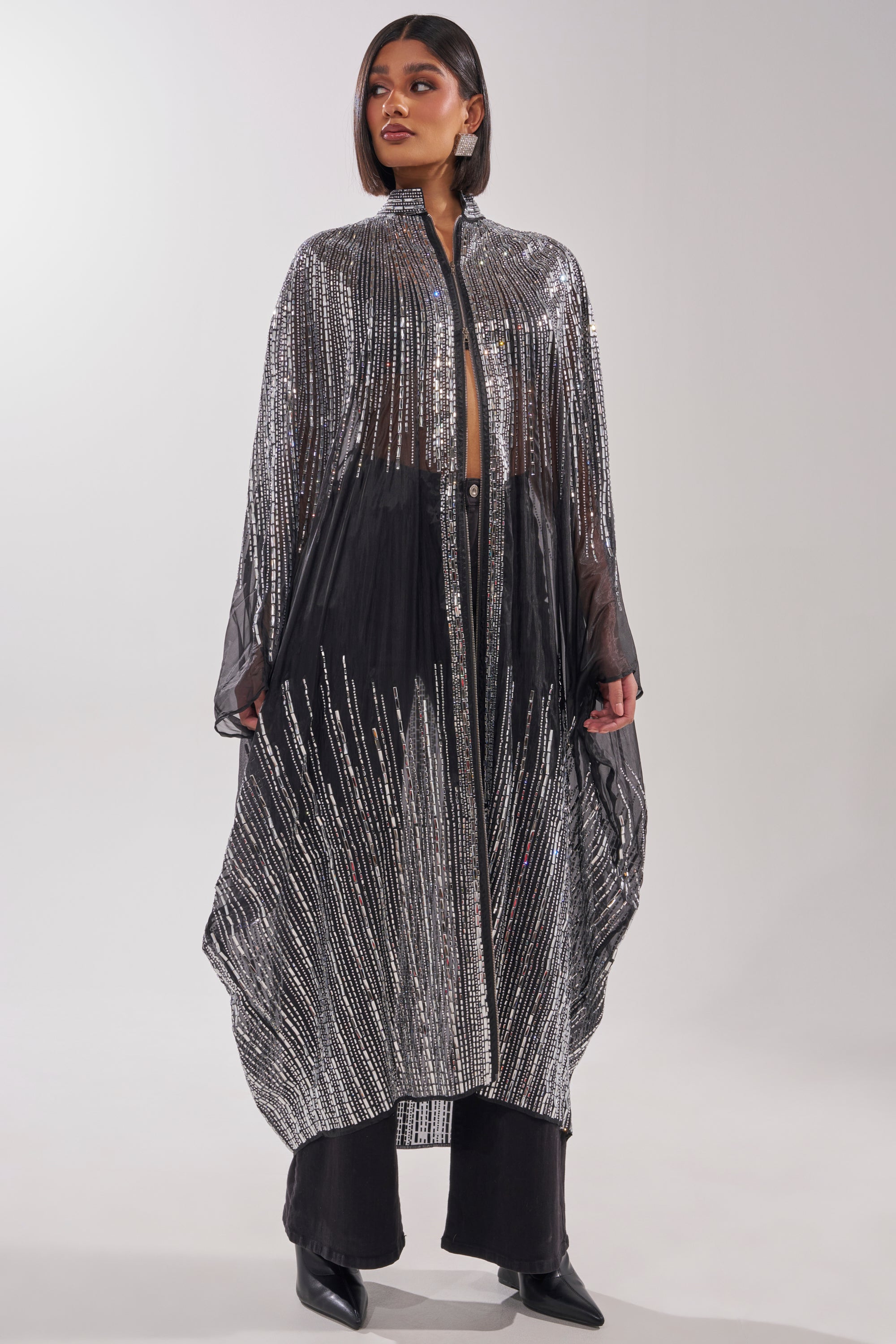 A woman poses confidently in the SPARKLE AND SHINE BLOUSE, a long, sheer black and silver sequin cape-style blouse with a high collar and vertical, diagonal shimmering patterns, styled over black pants and boots against a plain light background. 3