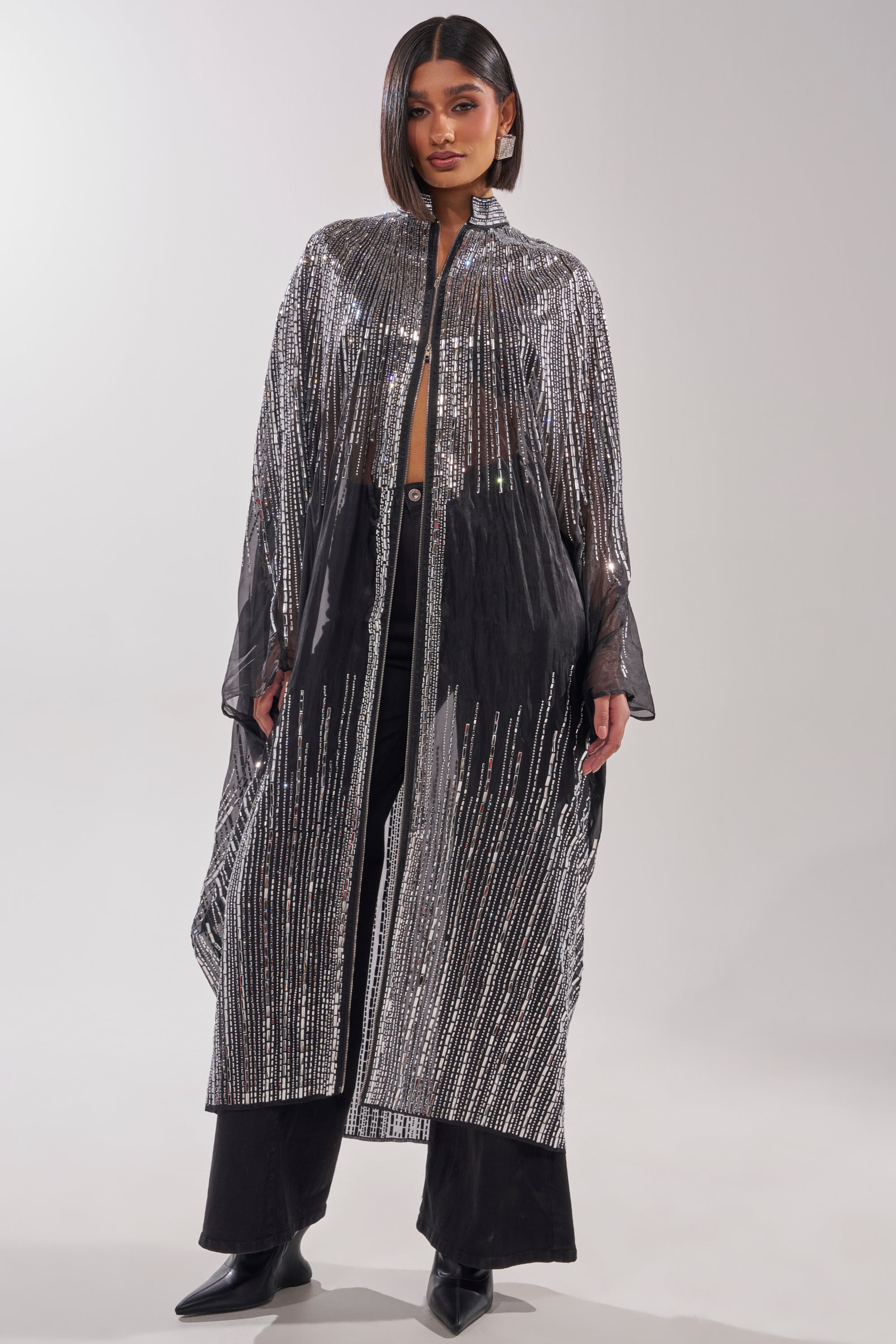 A woman wears the SPARKLE AND SHINE BLOUSE, a sheer, poncho-style black top with vertical silver sequins, paired with black pants and pointed-toe boots against a plain light background. 4