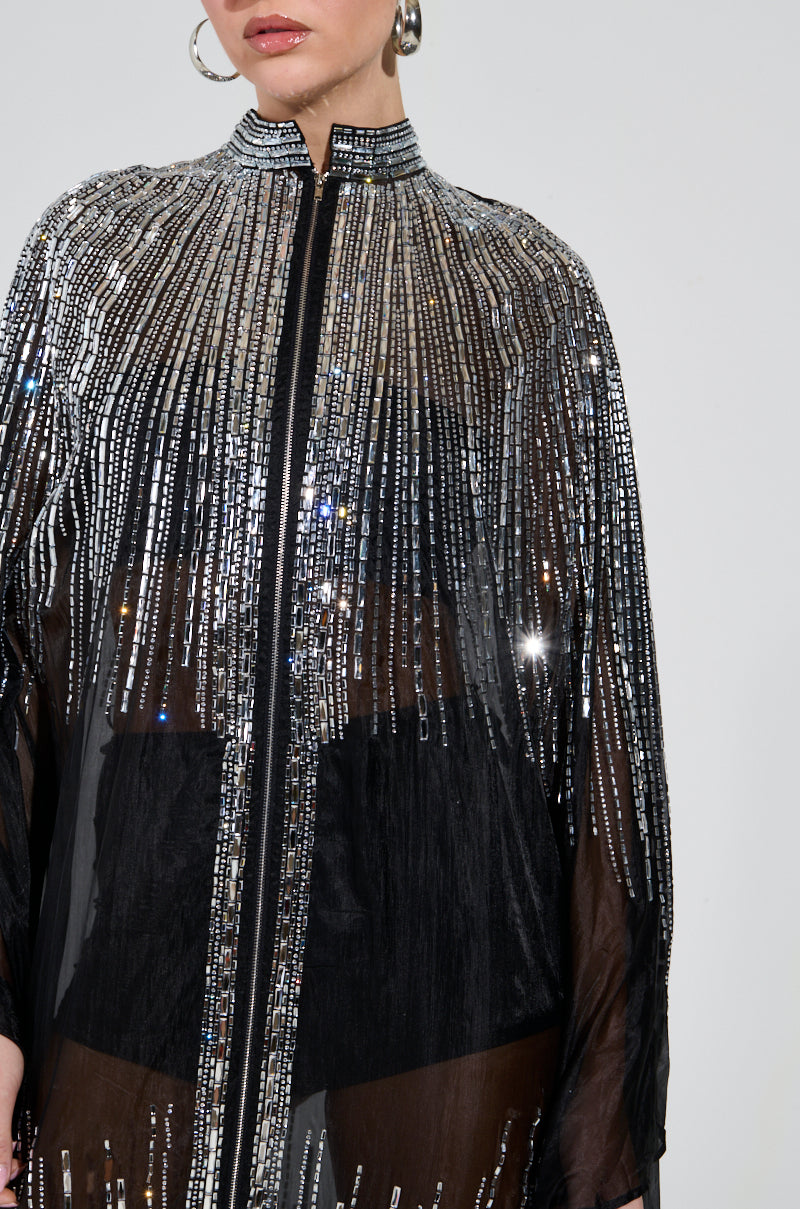 The SPARKLE AND SHINE BLOUSE is adorned with shimmering black and silver mirrored sequins, featuring long sleeves and a high collar. Its vertical lines create a dazzling effect, beautifully complemented by large hoop earrings against a plain, light-colored background.