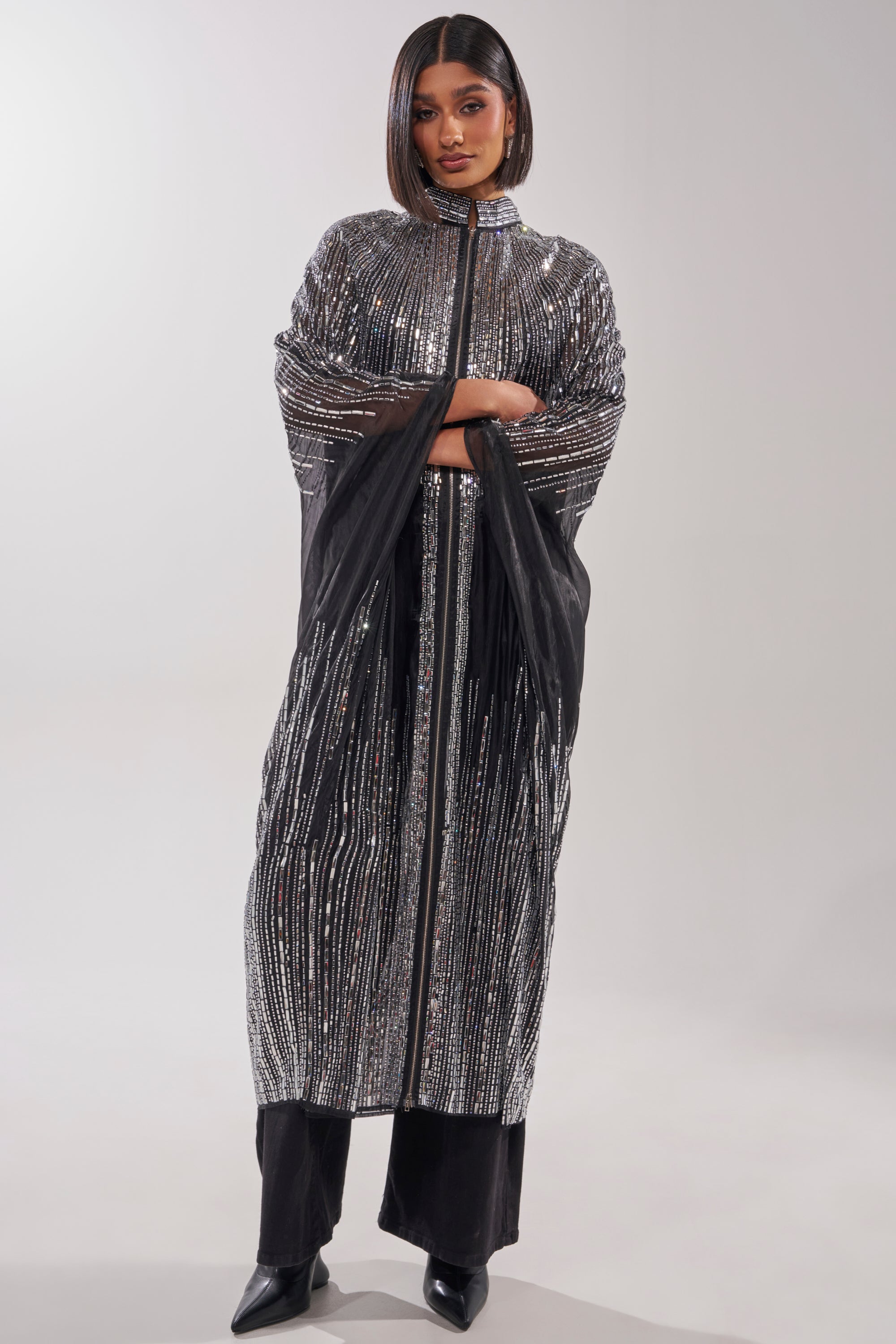 A woman stands with arms crossed, styled in the SPARKLE AND SHINE BLOUSE under a long, black and silver embellished abaya with vertical sparkling stripes, paired with black pants and pointed shoes against a plain light background. 5