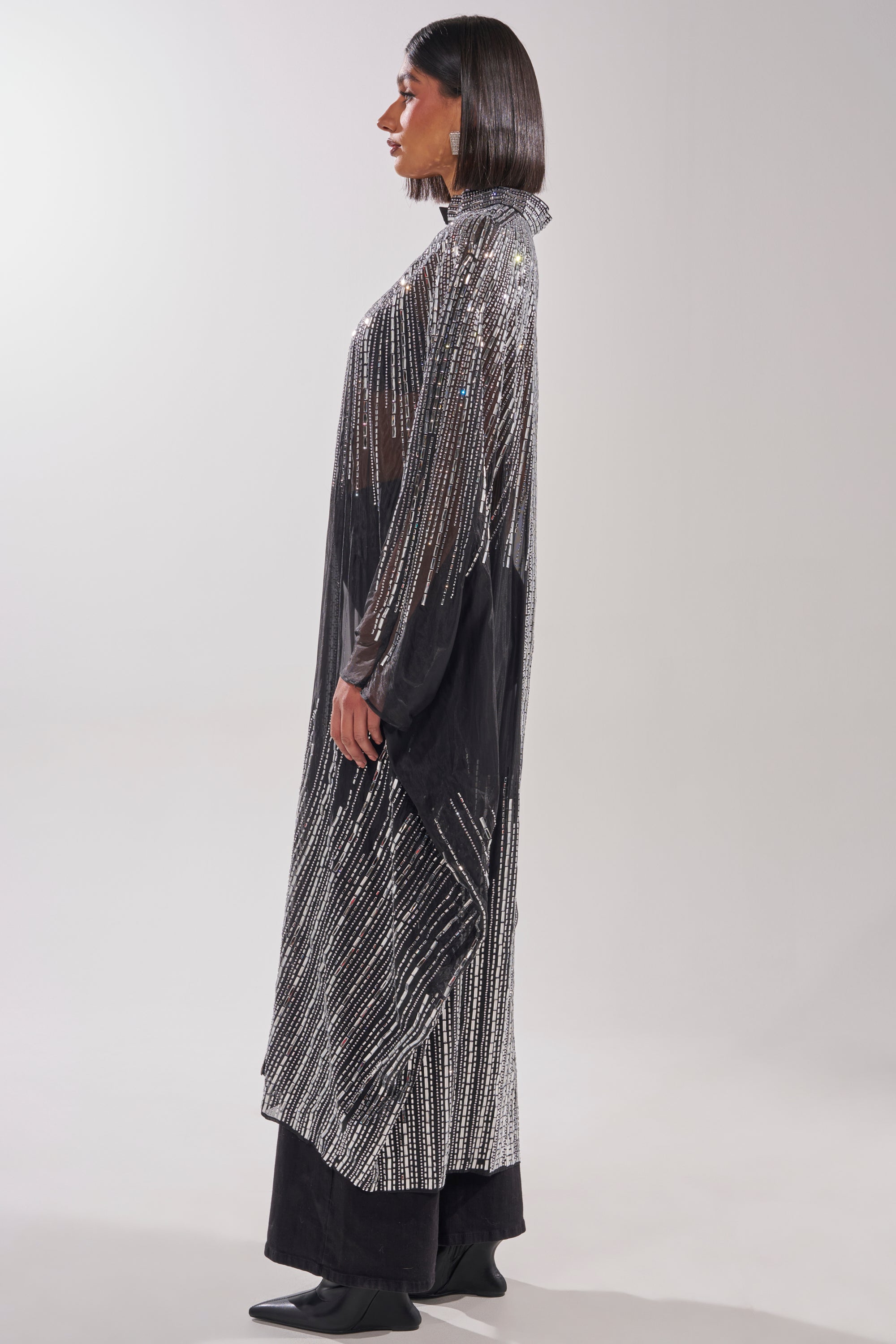 A woman stands in profile wearing the SPARKLE AND SHINE BLOUSE, a long, sheer black top with vertical silver sequin stripes, layered over black pants and boots against a plain, light-colored background. 6