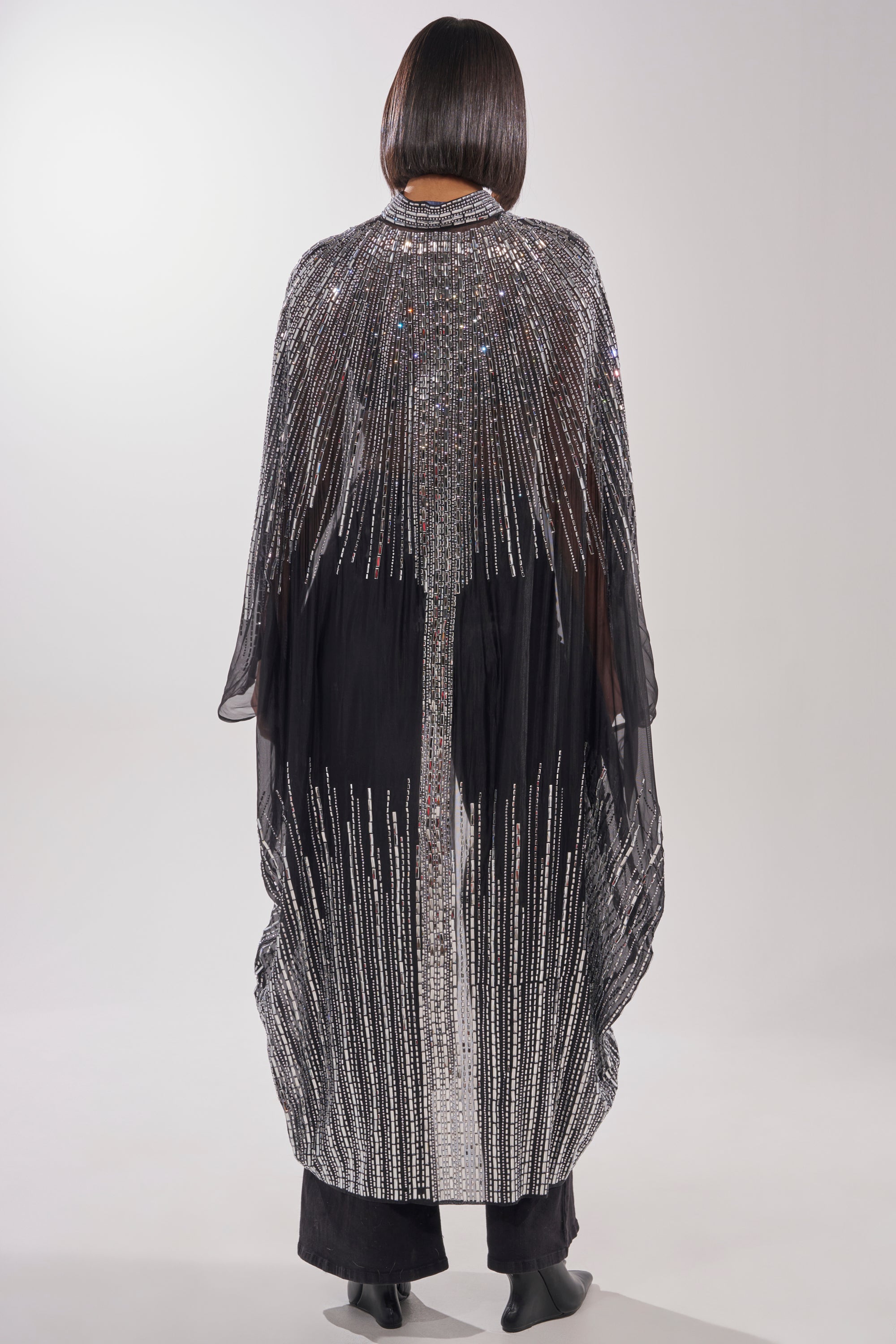A person stands facing away, wearing the SPARKLE AND SHINE BLOUSE—a long, black sheer top embellished with silver sequins in vertical lines for a radiant look. Their straight, chin-length hair is sleek and glossy. 7