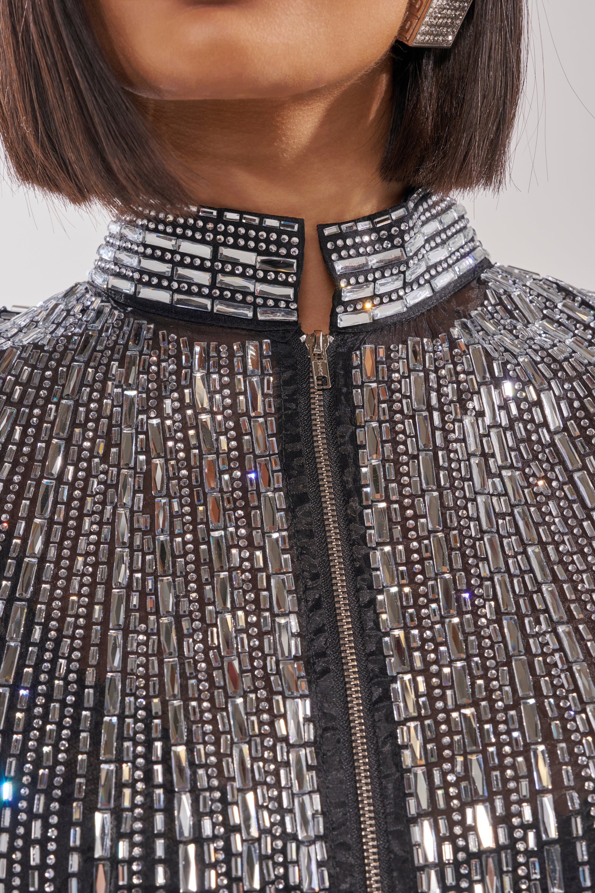 A close-up of a person wearing the SPARKLE AND SHINE BLOUSE, a zip-up high-collar jacket with rectangular and square silver sequins for a mosaic effect, paired with large square earrings. 8