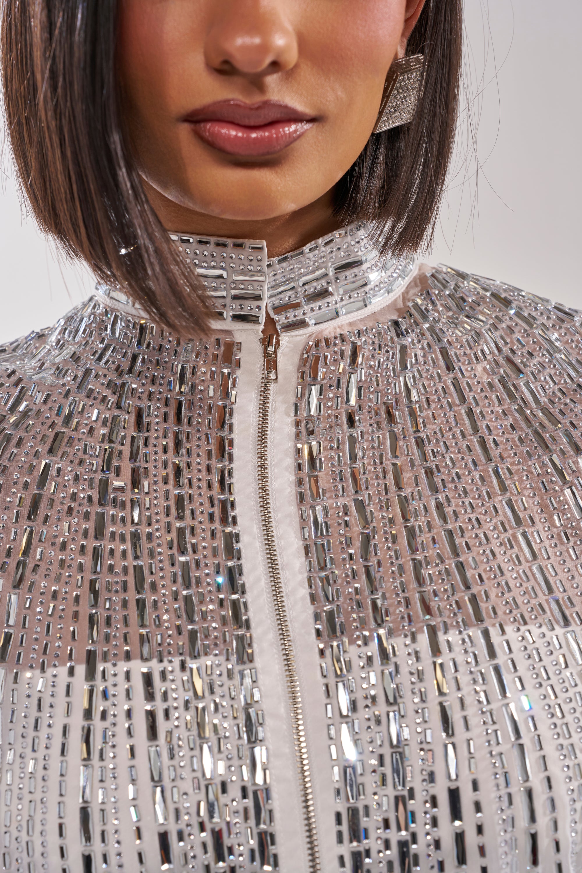 A woman with straight, shoulder-length dark hair wears the SPARKLE AND SHINE BLOUSE IN WHITE—a high-collared jacket covered in silver rectangular sequins with matching square earrings. The close-up highlights the blouse and her lower face. 9