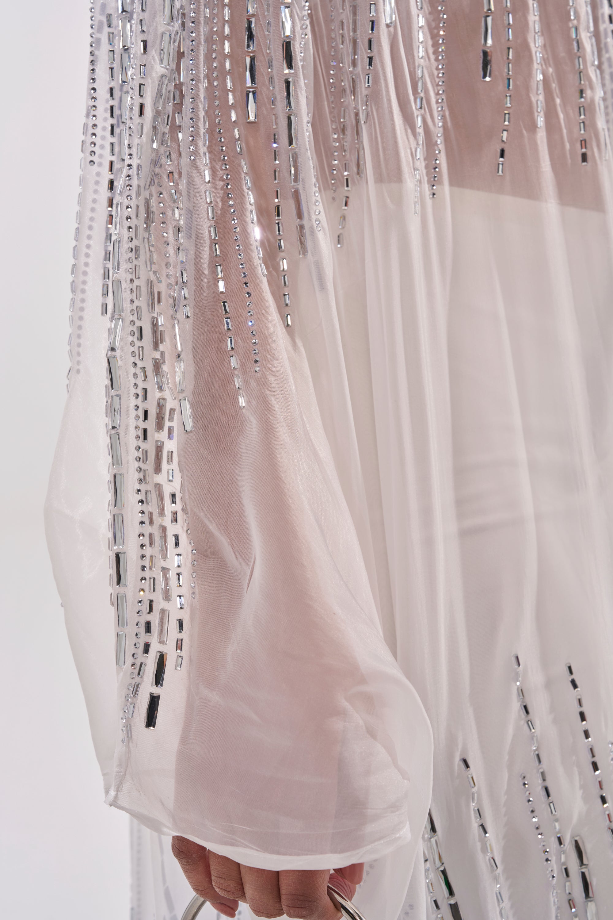 A close-up of the SPARKLE AND SHINE BLOUSE IN WHITE, featuring sheer nylon fabric with silver sequin embellishments and a hand gently holding the flowing, translucent material. 10