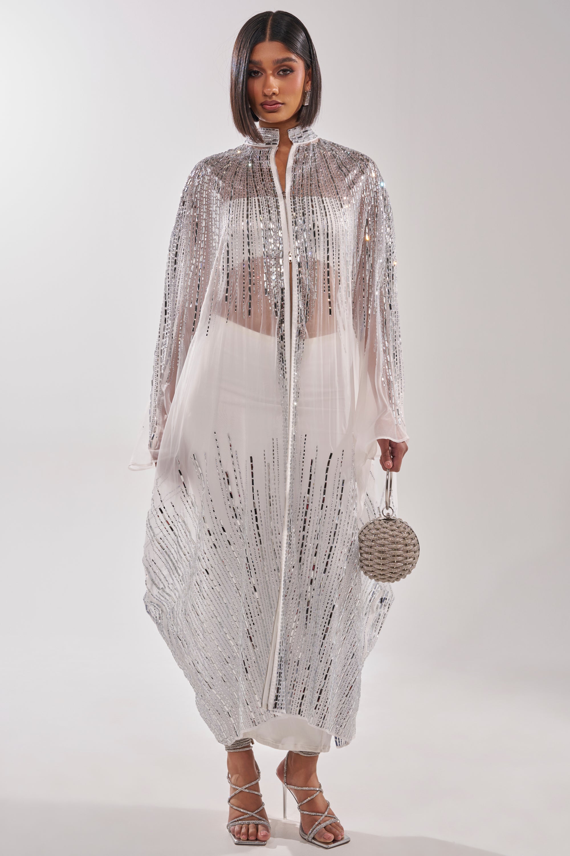 A woman wears the SPARKLE AND SHINE BLOUSE IN WHITE under a sheer, silver-sequined cape dress, paired with a round silver handbag and strappy silver heels, standing against a plain light background. 0