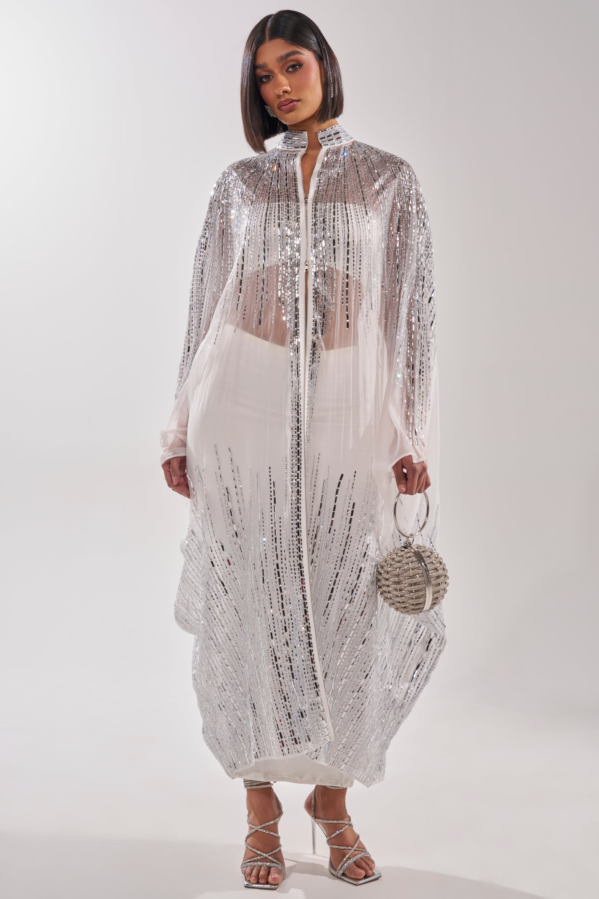 A woman exudes effortless glamour in the SPARKLE AND SHINE BLOUSE IN WHITE, styled as a long coat over a white outfit with strappy heels and a round silver bag, her sleek bob completing the chic look against a white background. 1