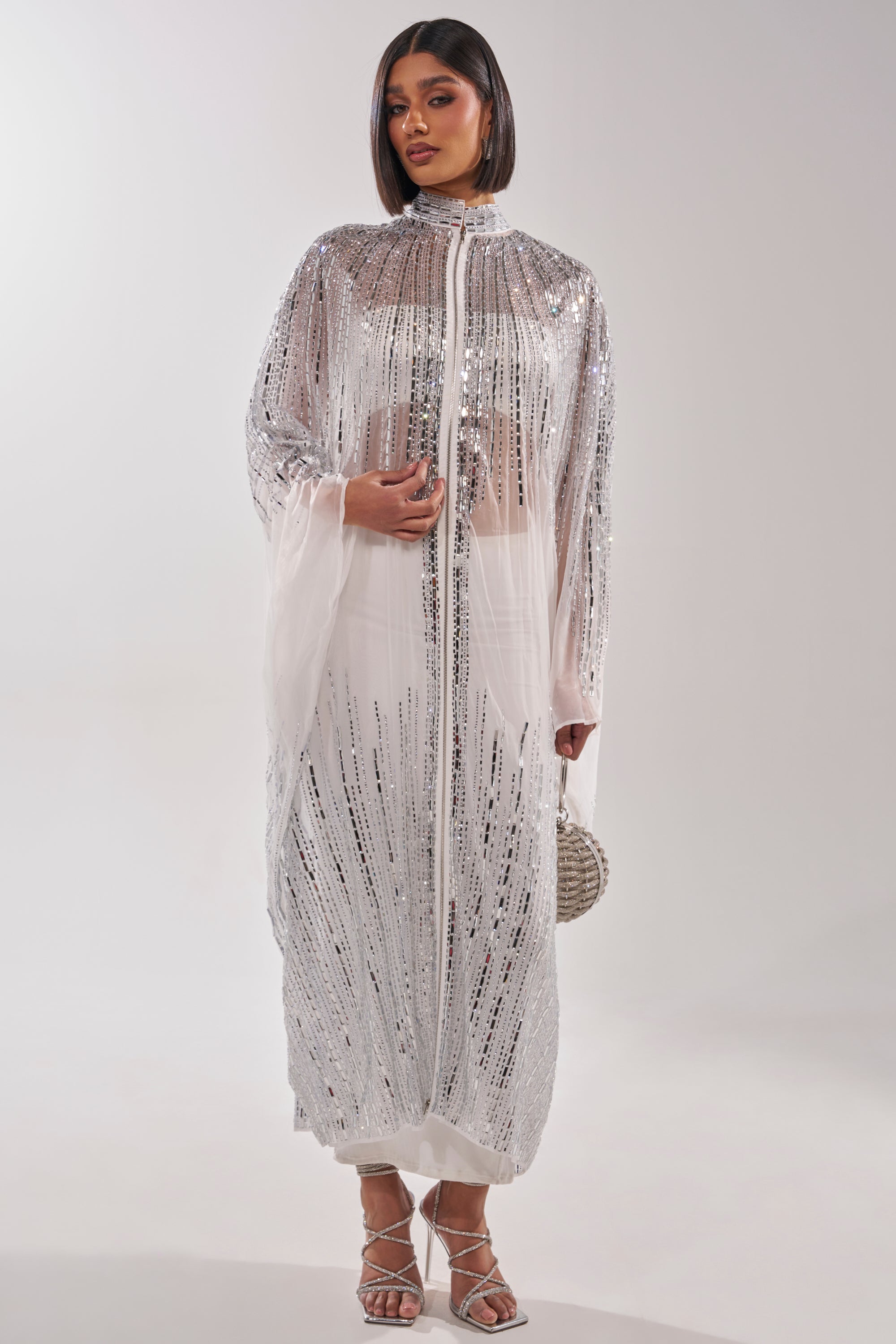 A woman models the SPARKLE AND SHINE BLOUSE IN WHITE over a white dress, holding a small round silver bag. She completes the look with strappy heels and wears her straight, shoulder-length hair down against a plain light background. 2