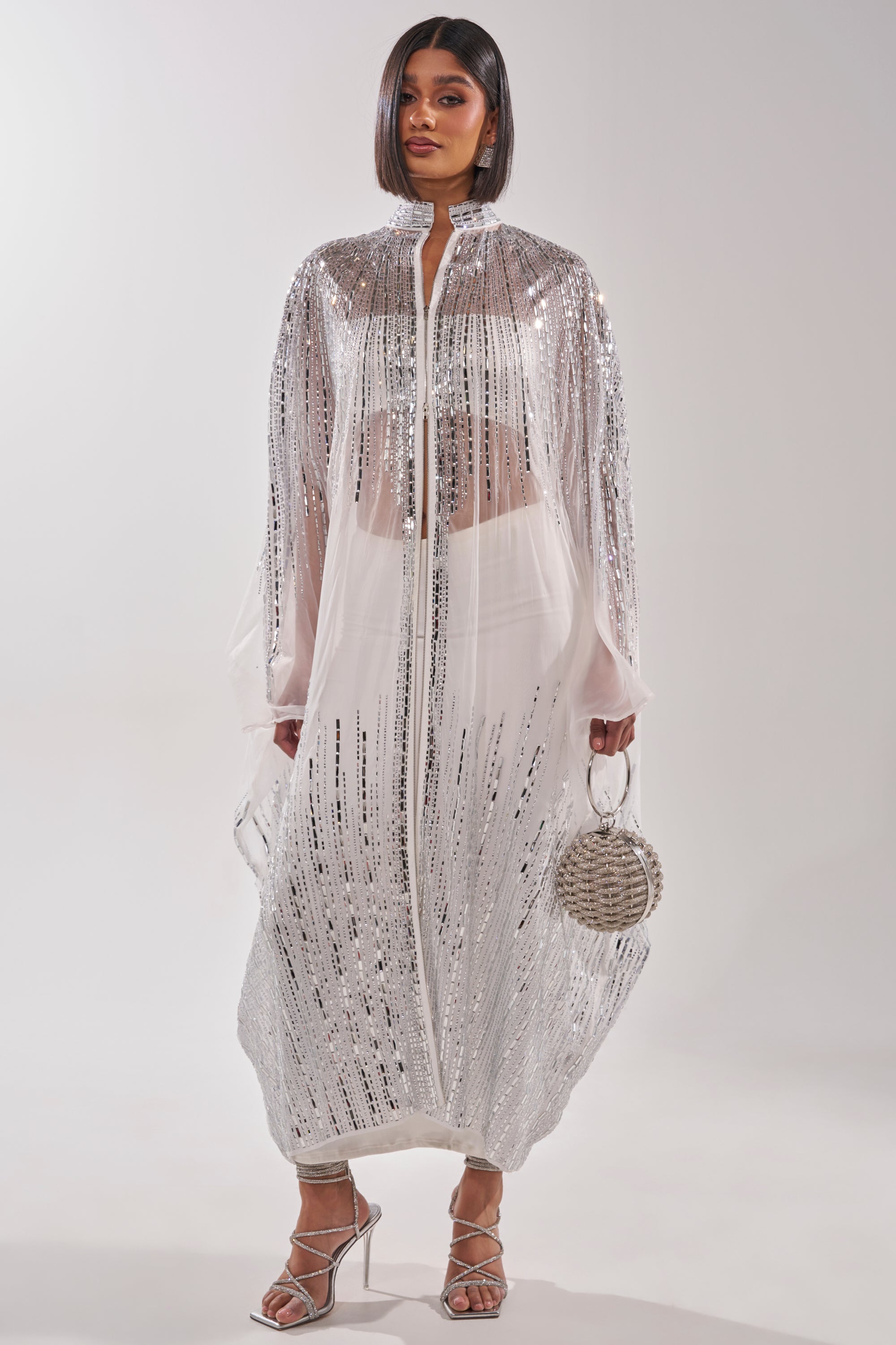 A woman poses against a white backdrop in a long, sheer, silver sequined coat over the SPARKLE AND SHINE BLOUSE IN WHITE, paired with strappy silver heels and a round silver handbag. Her hair is sleek and straight. 4
