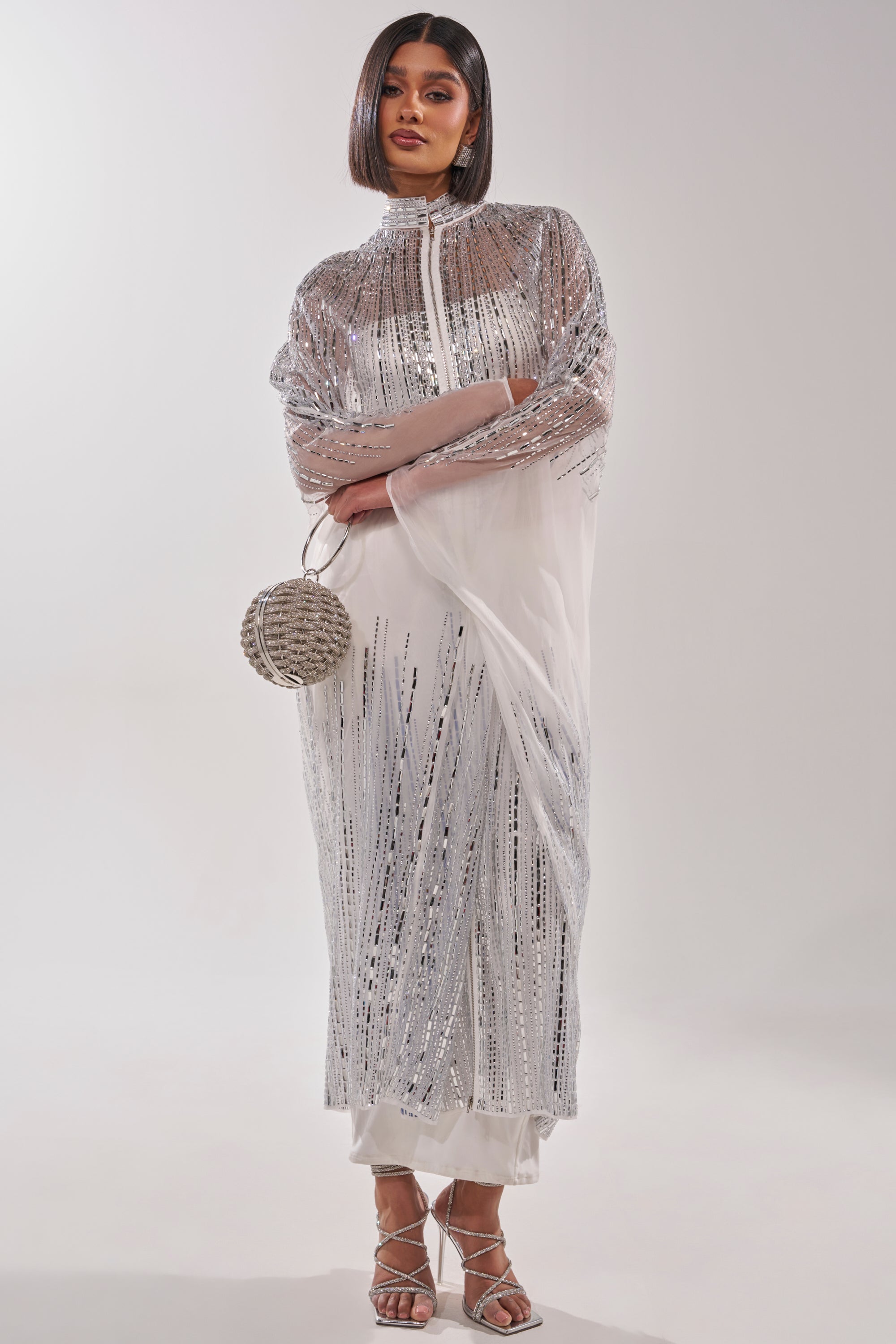 A woman wears the SPARKLE AND SHINE BLOUSE IN WHITE, a floor-length white sequin blouse dress with silver sequins and sheer sleeves, styled with loose pants, strappy heels, and a round silver purse against a plain white background. 5
