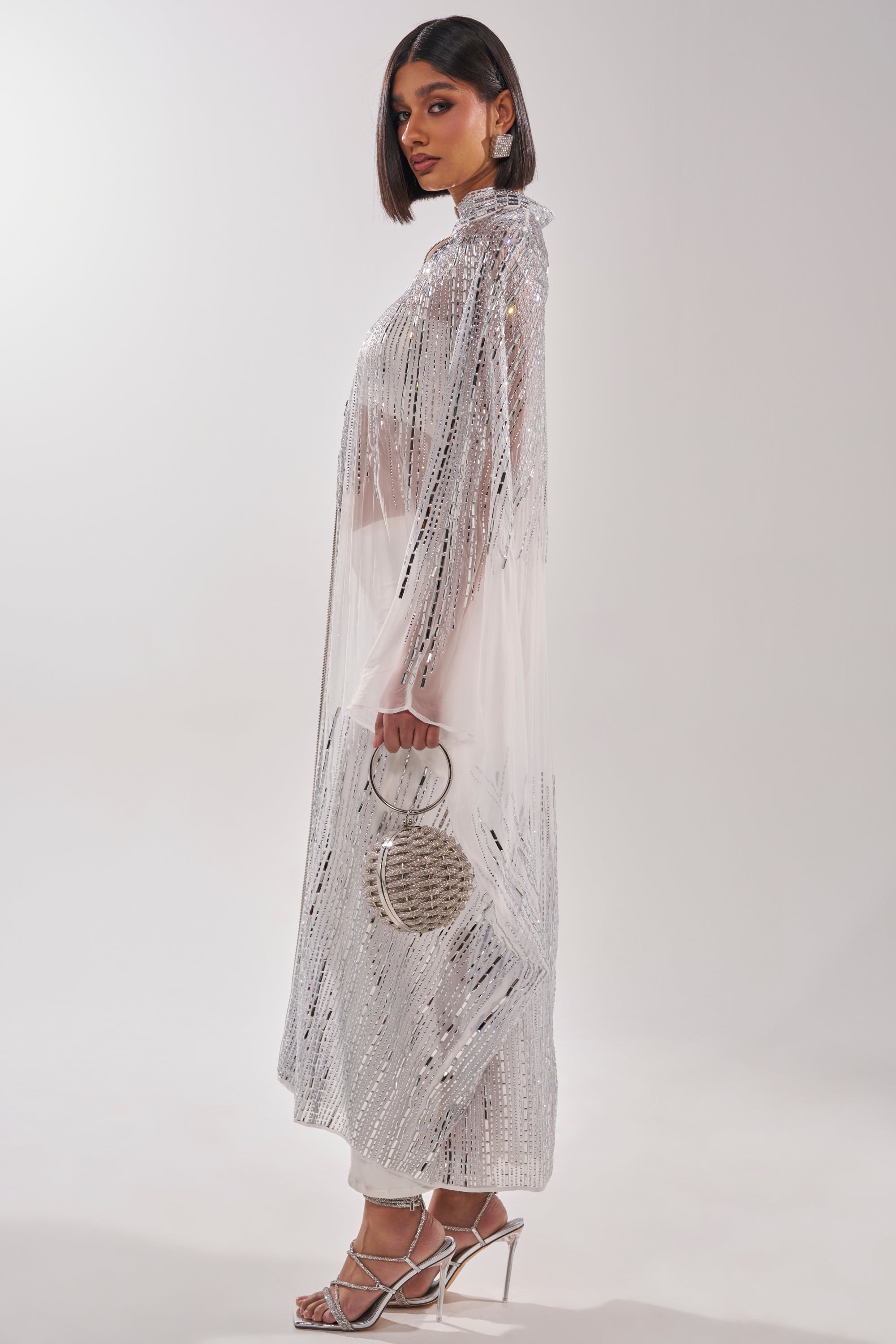 A woman in a sheer, sparkling white Sparkle and Shine Blouse with a high collar stands sideways, holding a round silver handbag. She wears strappy silver heels and has a sleek shoulder-length hairstyle against a plain background. 7