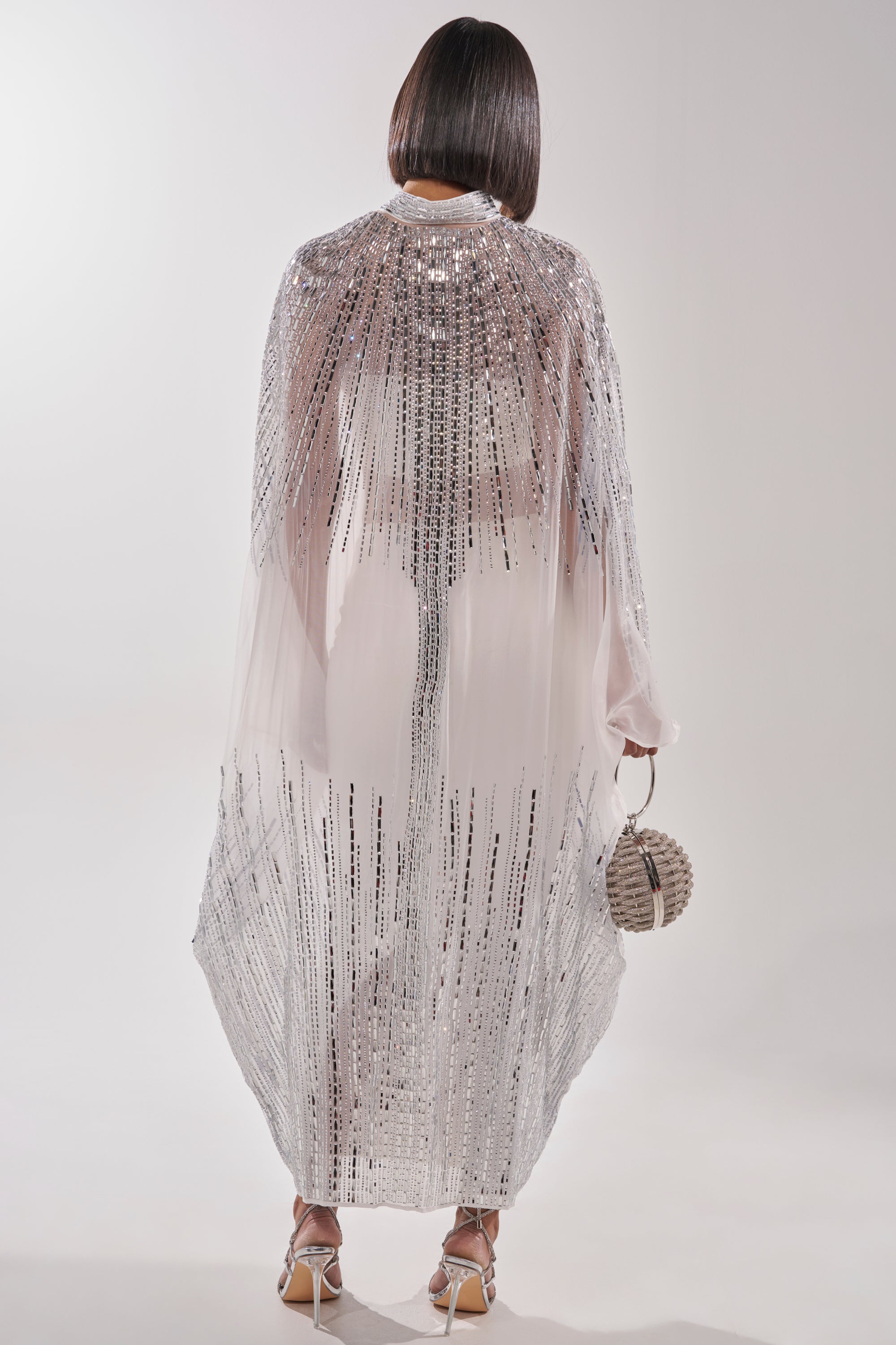 A person with a sleek bob faces away, wearing the SPARKLE AND SHINE BLOUSE IN WHITE paired with a sheer, sequin-detailed cape, white dress, round beaded handbag, and strappy heels against a plain white background. 8