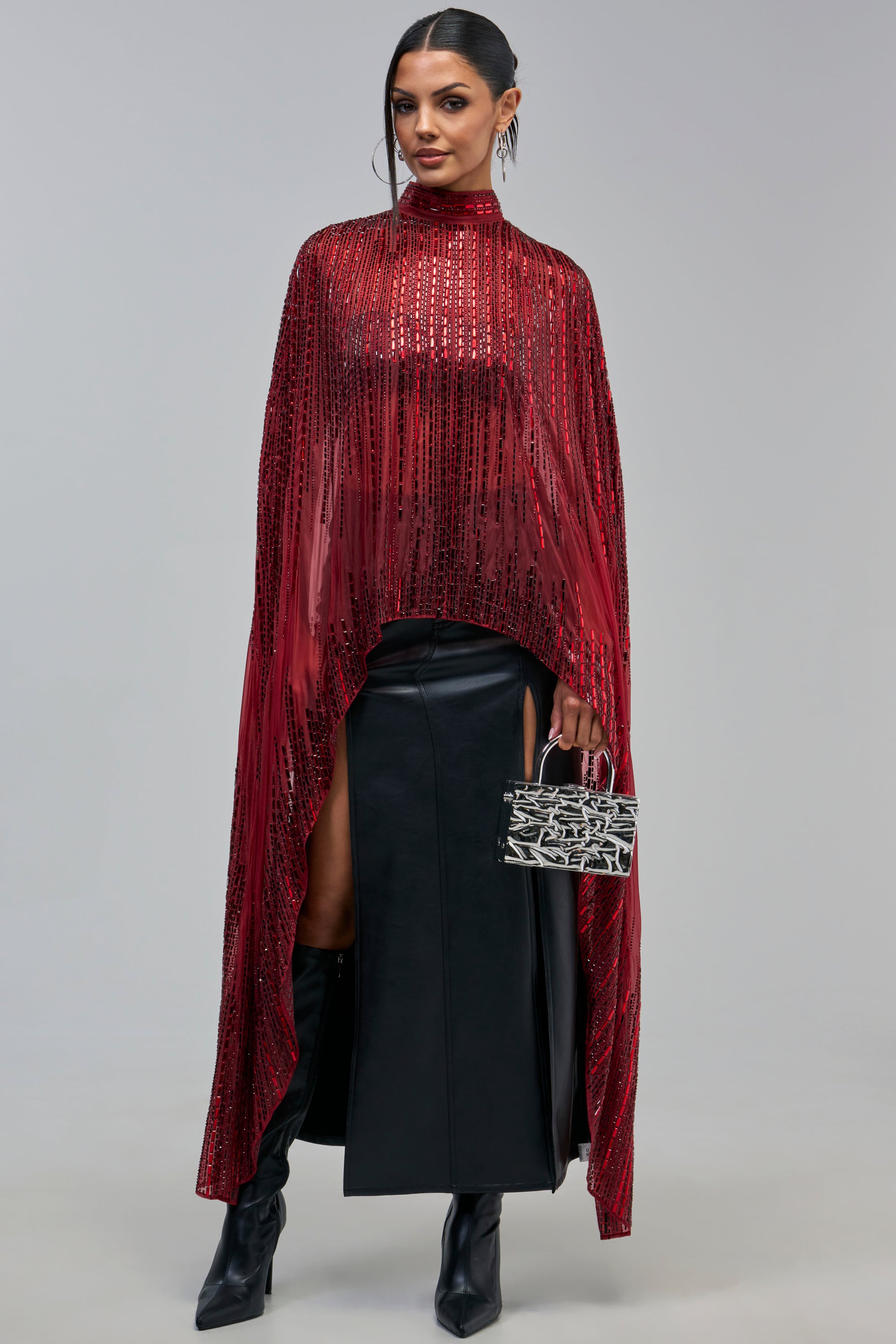 A person wears a DRIPPING IN RHINESTONES CAPE IN BURGUNDY over a black skirt and knee-high boots, holding a small silver and black handbag for evening styling.