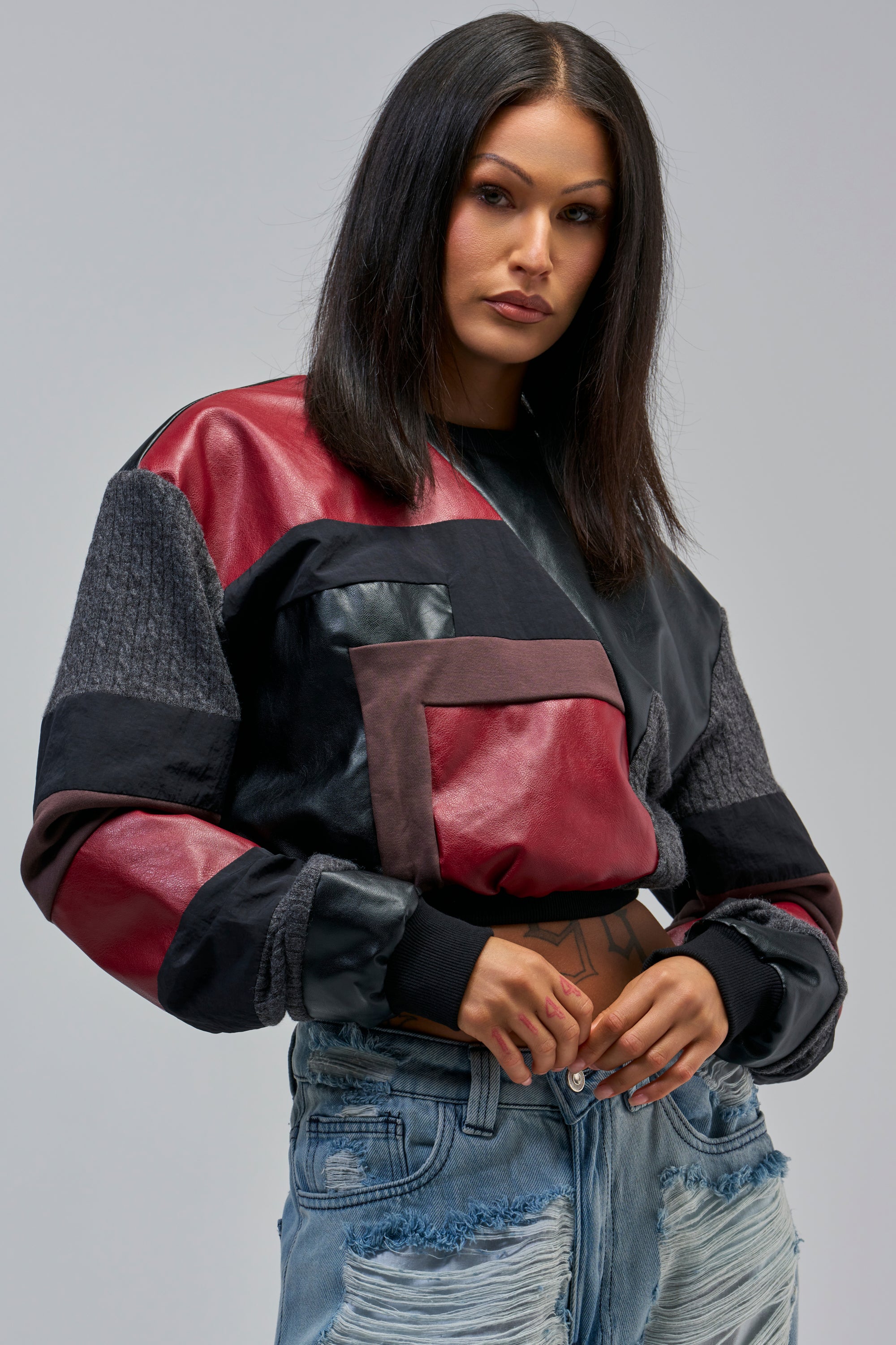 Woman with straight dark hair wears the PATCH FIX CROPPED SWEATSHIRT IN BLACK MULTI and light blue ripped jeans, posing with one hand on her waist against a plain gray background.