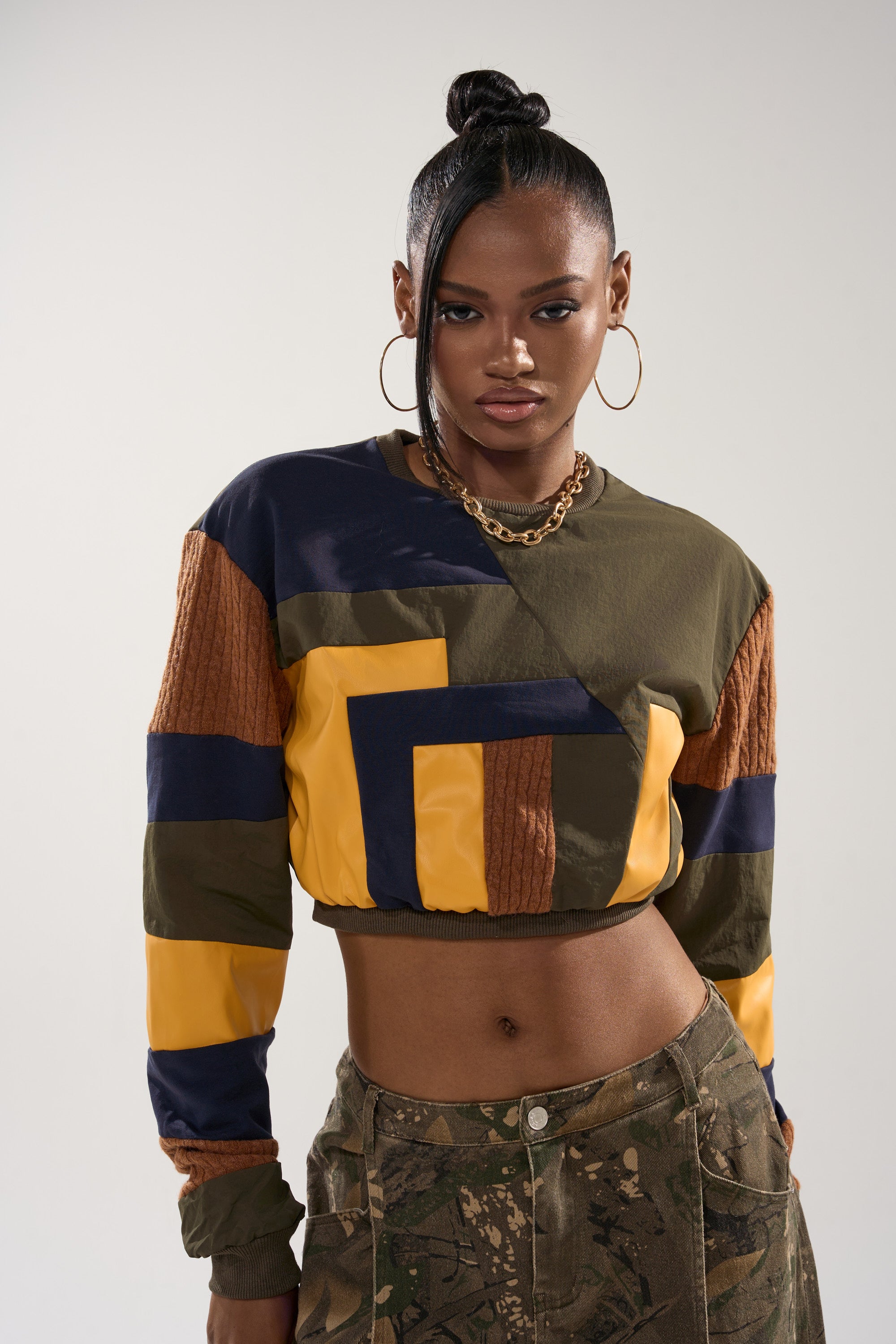 A woman with a high bun wears the DORA COLOR BLOCKED CROP SWEATSHIRT in olive, yellow, navy, and brown, paired with corduroy camo-pattern pants and gold hoop earrings, standing against a plain background.