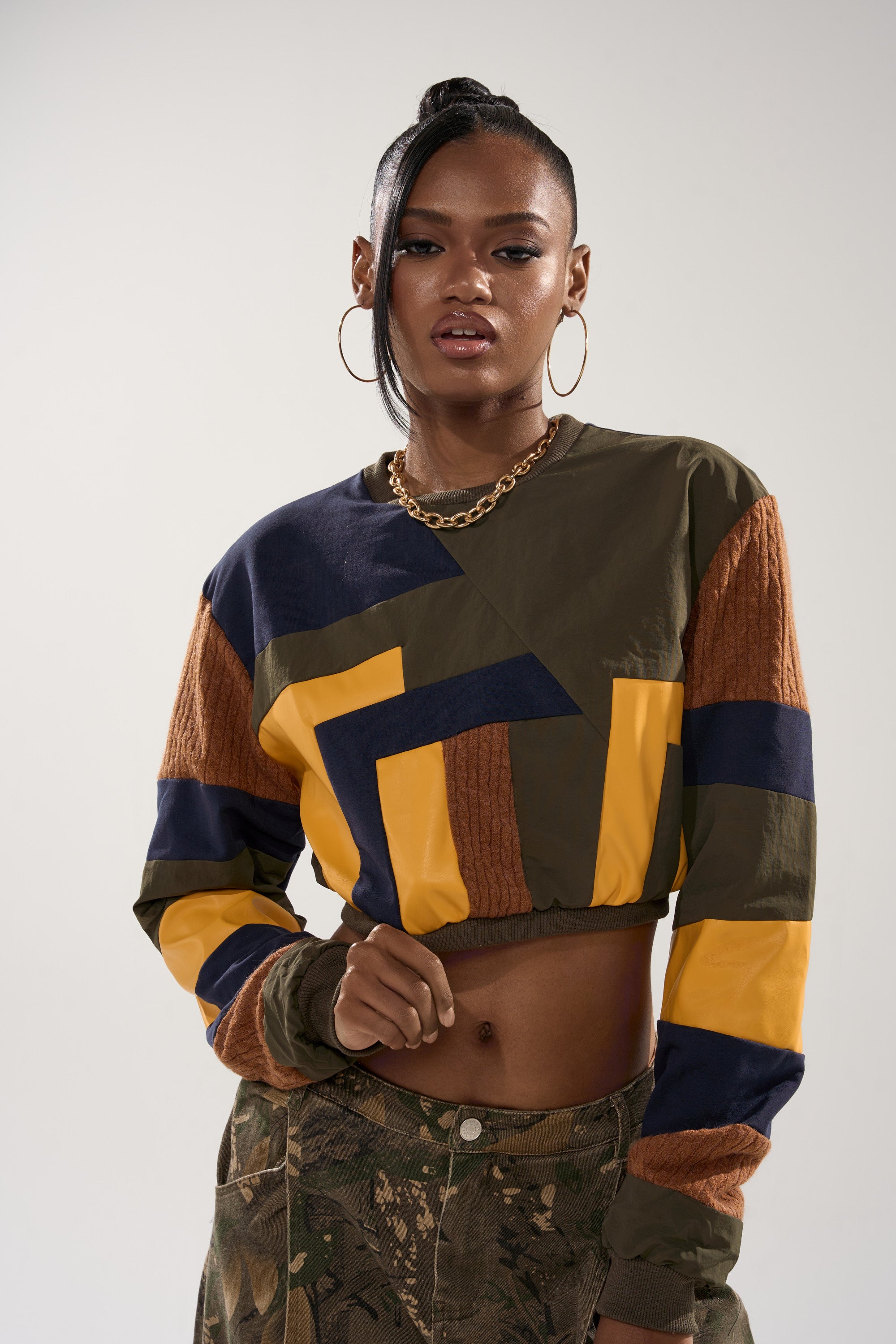 A woman poses confidently in the DORA COLOR BLOCKED CROP SWEATSHIRT, featuring yellow, brown, navy, and olive panels, paired with camouflage pants and a chunky gold chain necklace against a plain light background.