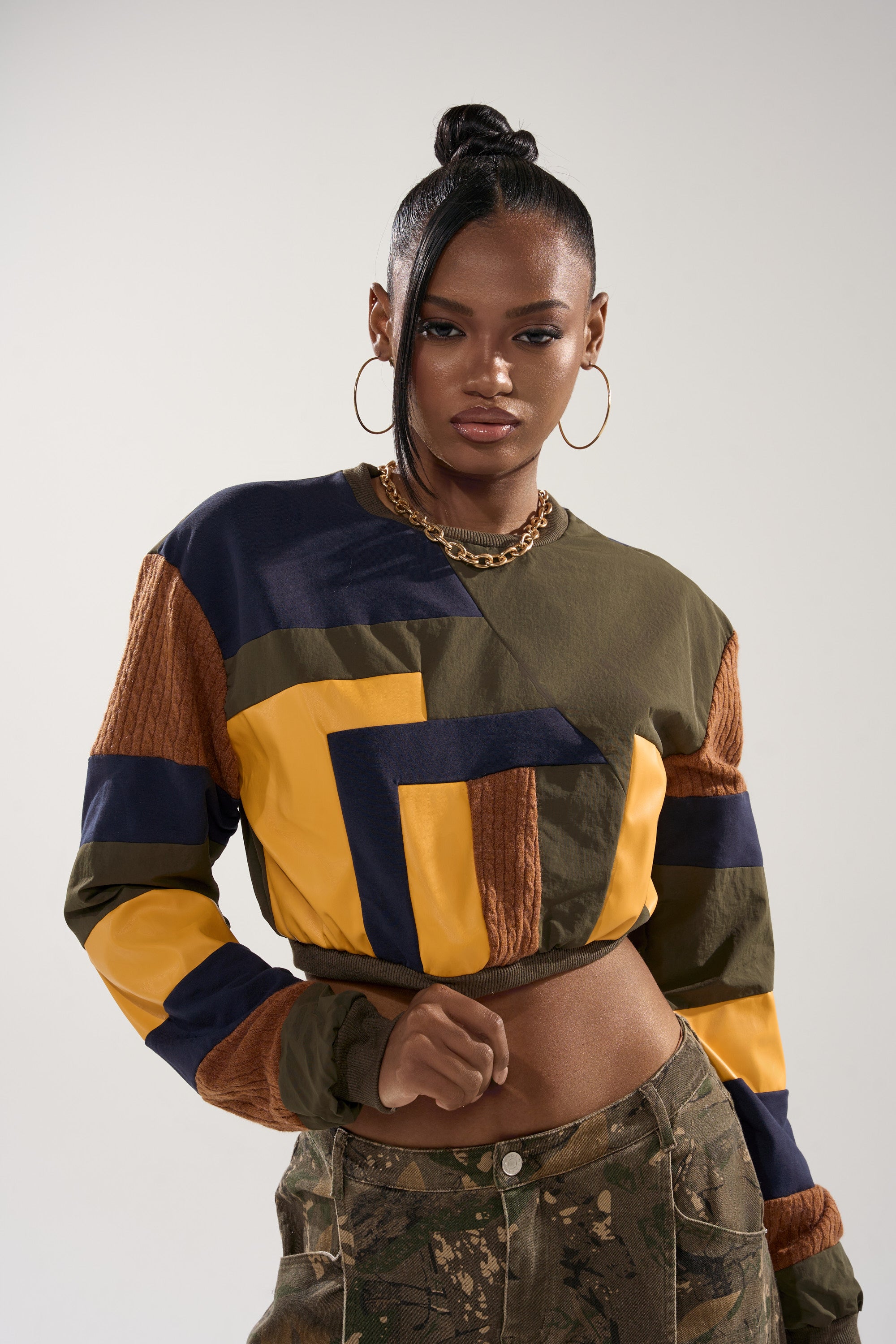 A woman with a high bun wears the DORA COLOR BLOCKED CROP SWEATSHIRT in yellow, navy, olive, and brown, paired with camouflage pants. She accessorizes with hoop earrings and a chunky chain necklace.