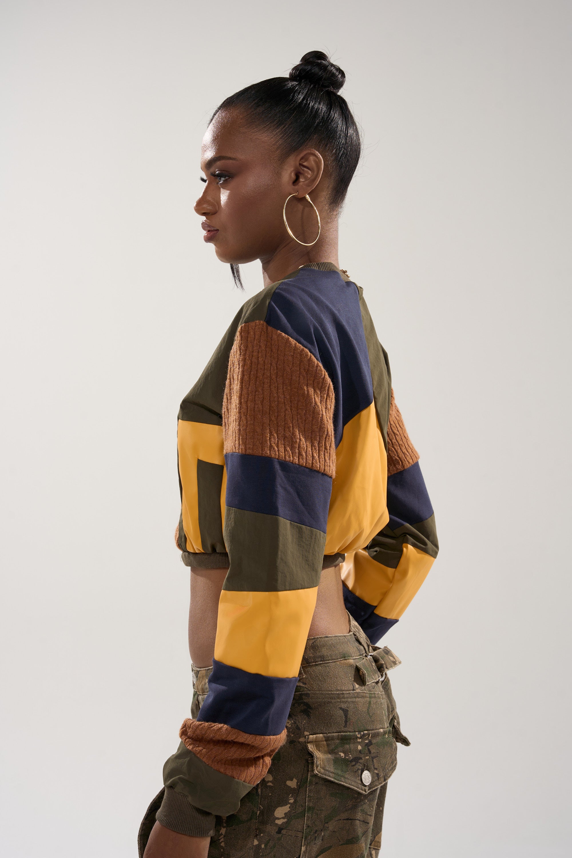 Wearing hoop earrings and a high bun, a person models the DORA COLOR BLOCKED CROP SWEATSHIRT in yellow, navy, brown, and olive green with camouflage pants, standing in profile against a light background.
