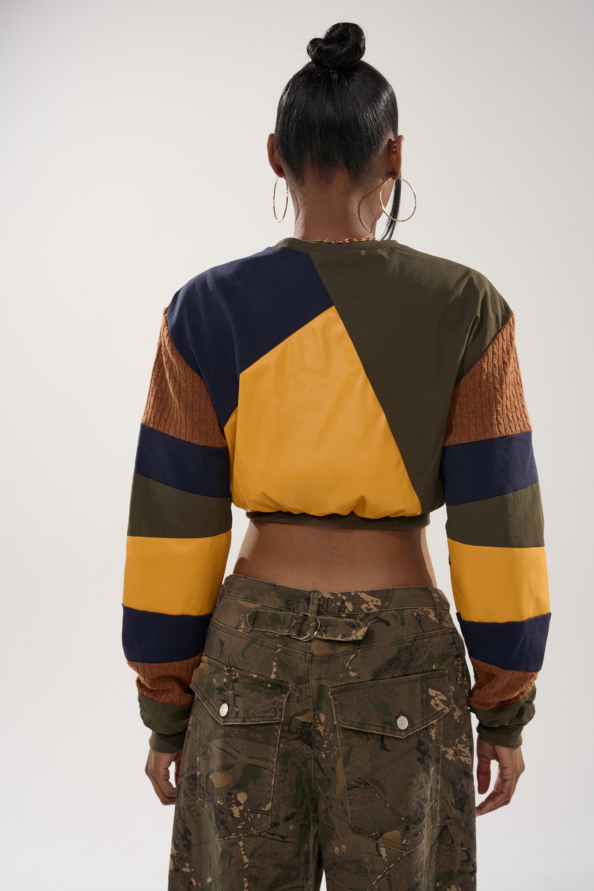 Wearing the DORA COLOR BLOCKED CROP SWEATSHIRT, a person with a bun, large hoop earrings, and streetwear style faces away from the camera, paired with camo pants, against a plain background.