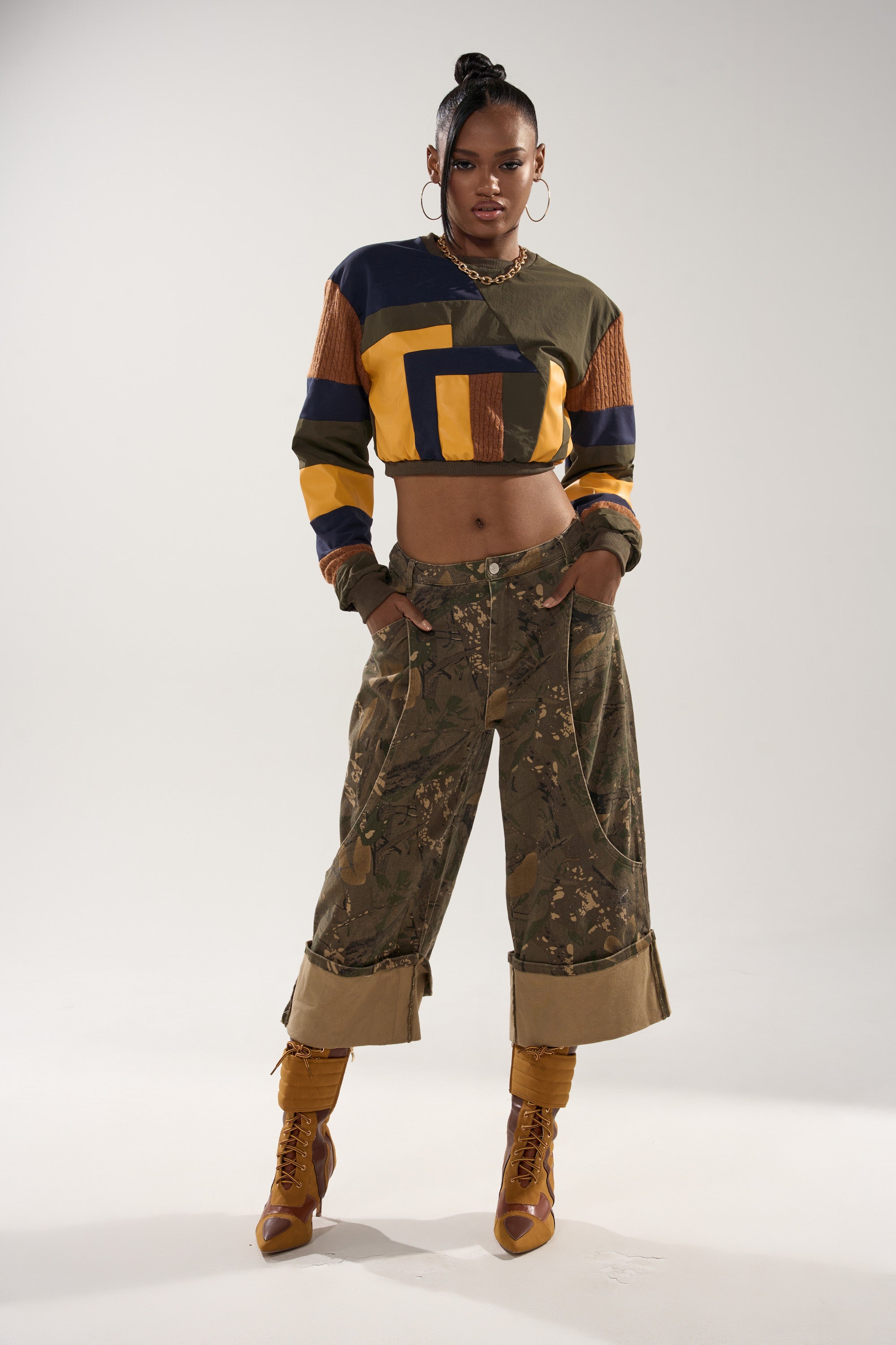 A woman stands confidently in a studio wearing the DORA COLOR BLOCKED CROP SWEATSHIRT, camo pants, and mustard lace-up boots. Her high ponytail and hands-in-pockets pose exude streetwear flair.