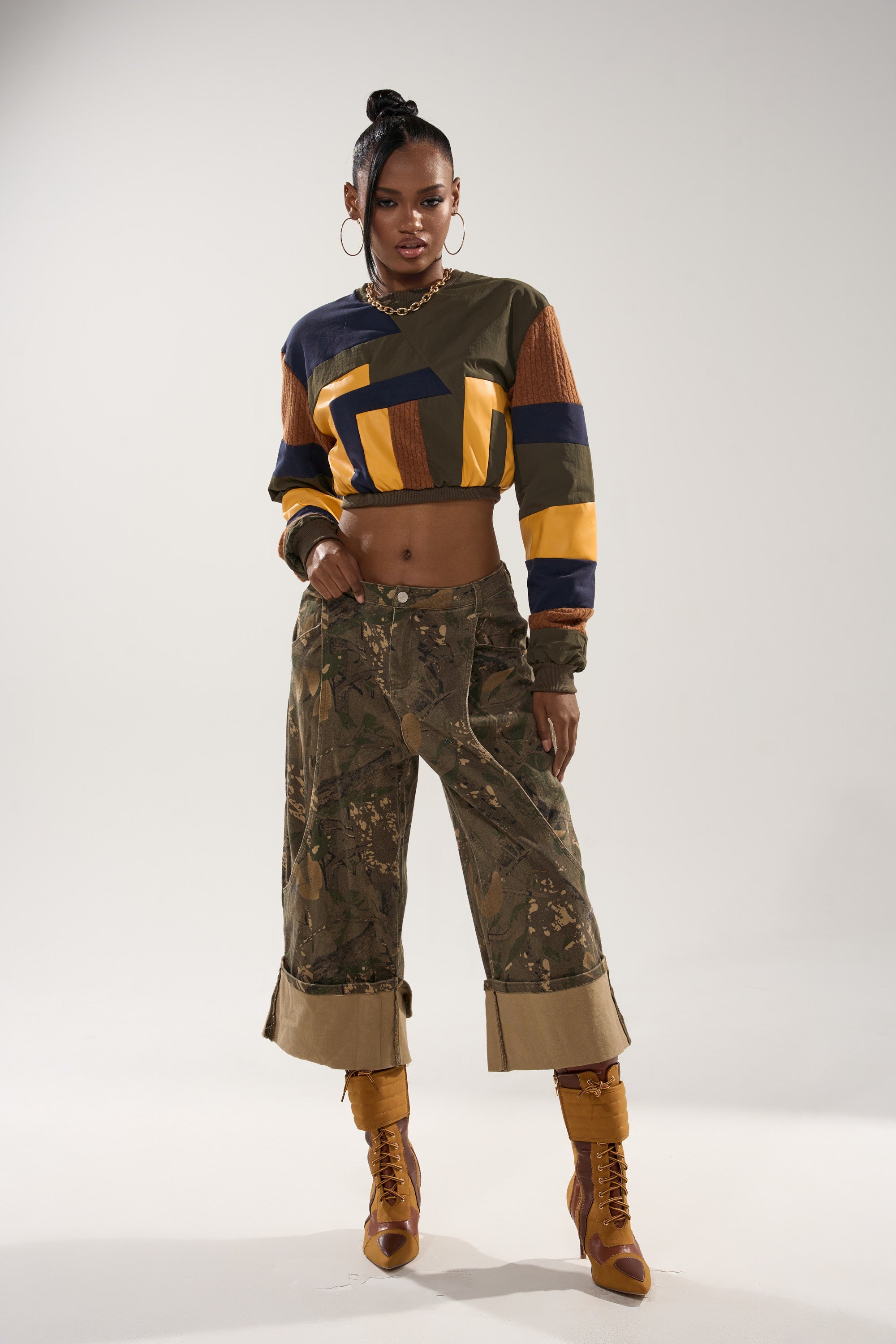 A woman stands confidently in the DORA COLOR BLOCKED CROP SWEATSHIRT, camo wide-leg rolled-cuff pants, tan heeled boots, a chunky gold necklace, and large hoop earrings against a plain background.