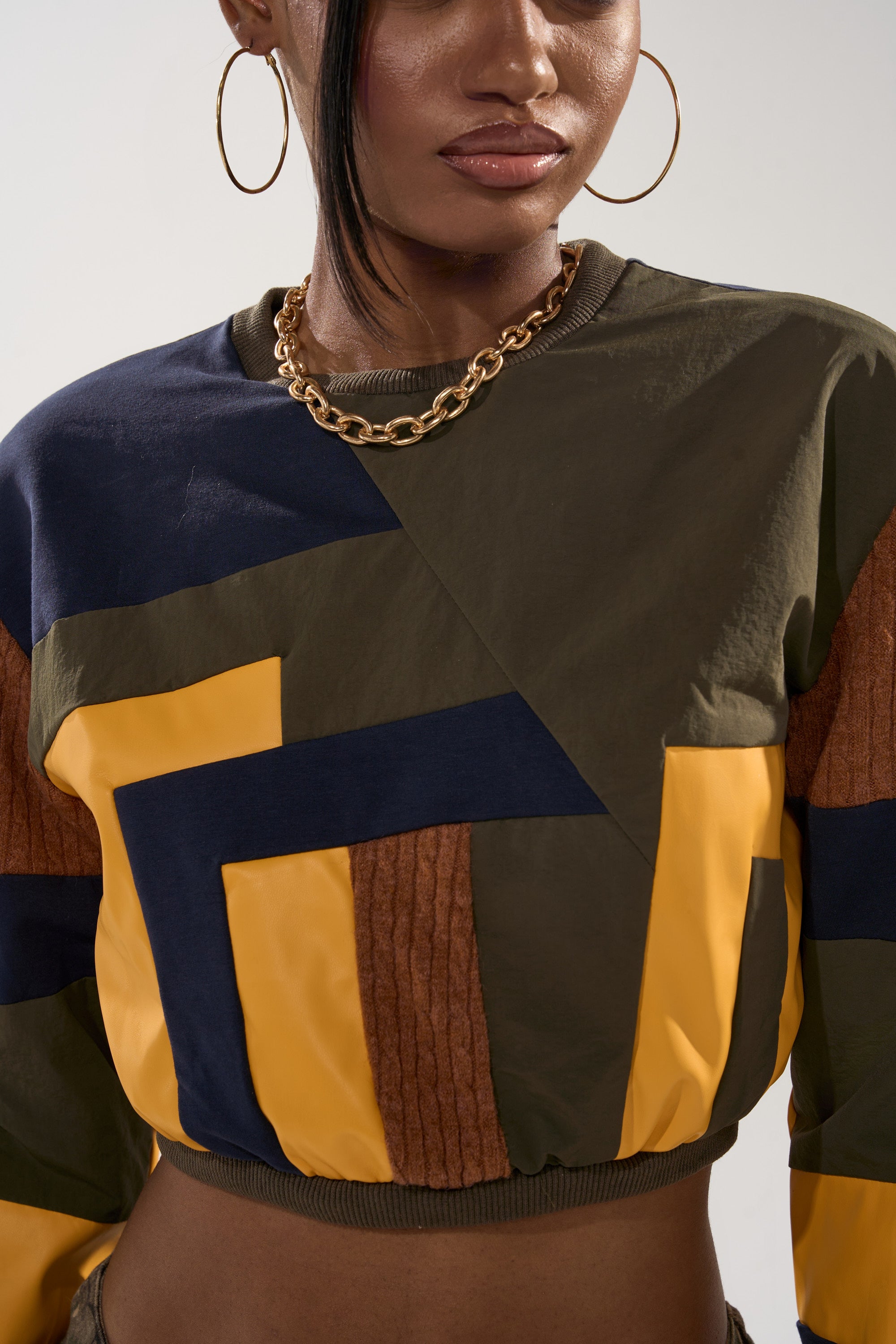 A woman wears the DORA COLOR BLOCKED CROP SWEATSHIRT with geometric yellow, navy, green, and brown patterns, styled with gold hoop earrings and a chunky gold chain for a streetwear look cropped from shoulders to midriff.