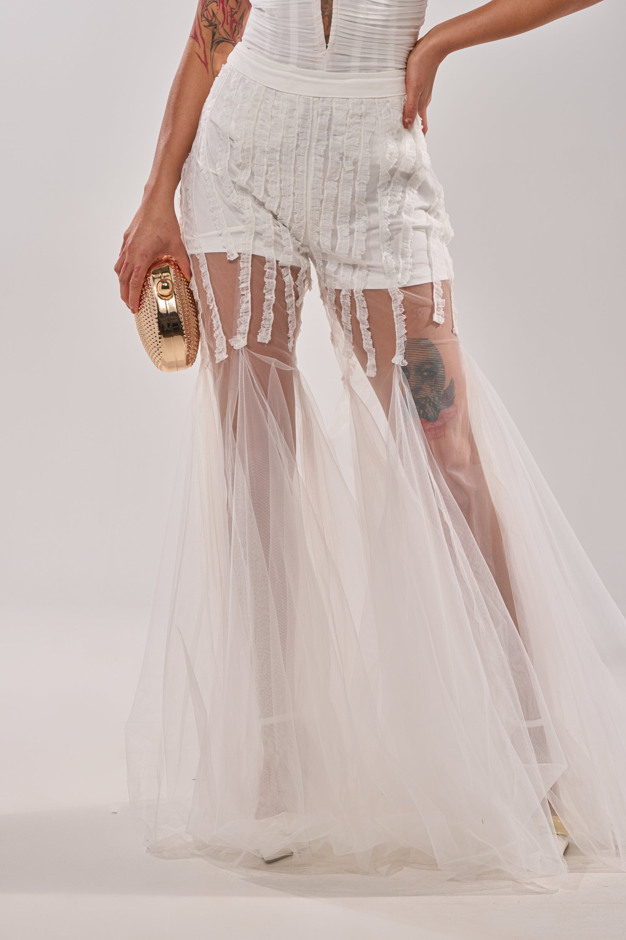 Wearing the EYE FOR MY EYES ON YOUR MESH FLARE PANT, this person styles a chic white look with sheer panels and a textured gold clutch. Visible tattoos add edge, making a bold statement against the plain background. 0