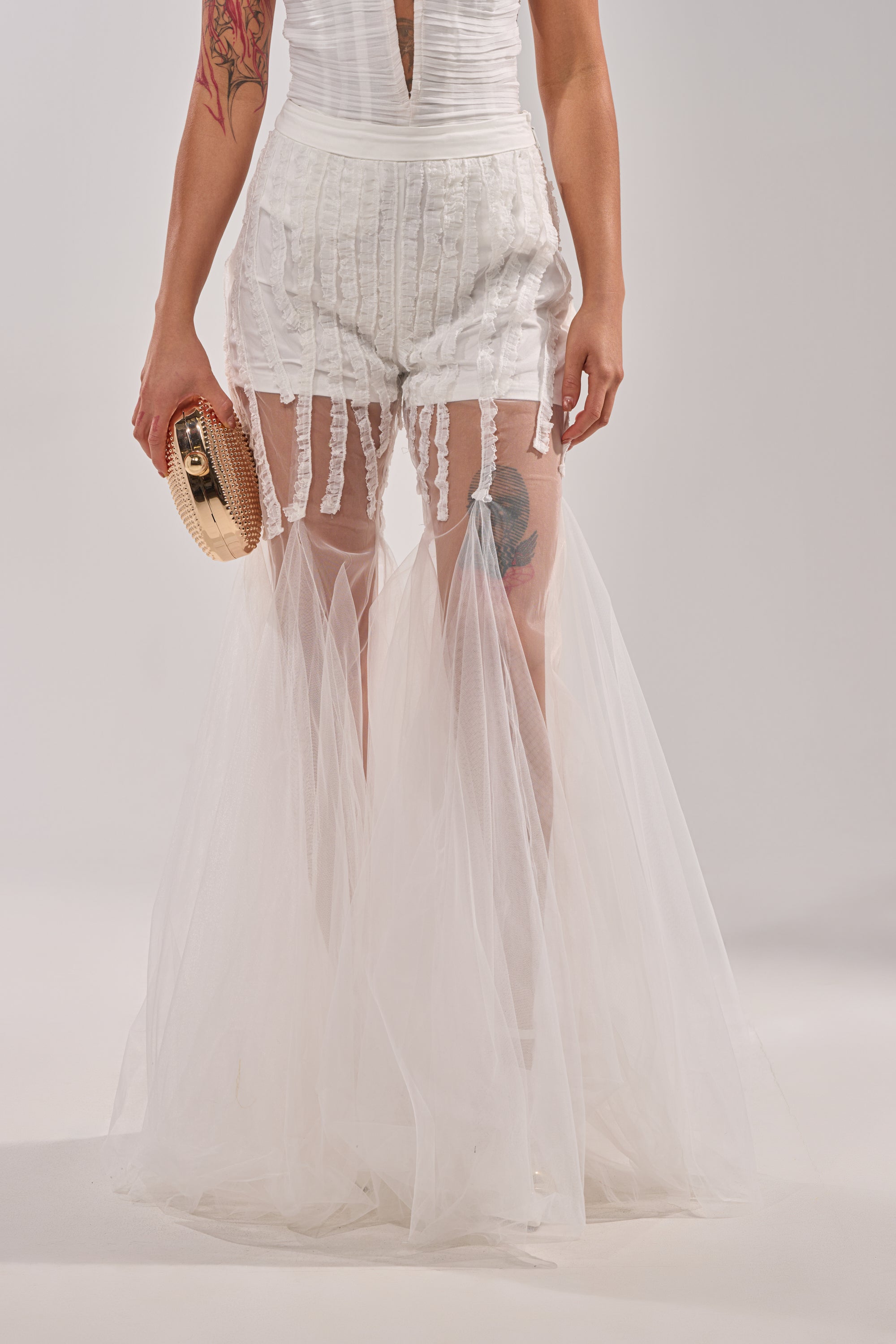 Wearing EYE FOR MY EYES ON YOUR MESH FLARE PANT—white, textured sheer mesh with ruffled flare legs for a skirt effect—paired with a small gold clutch and showcasing an upper thigh tattoo, this editorial look turns heads. 1