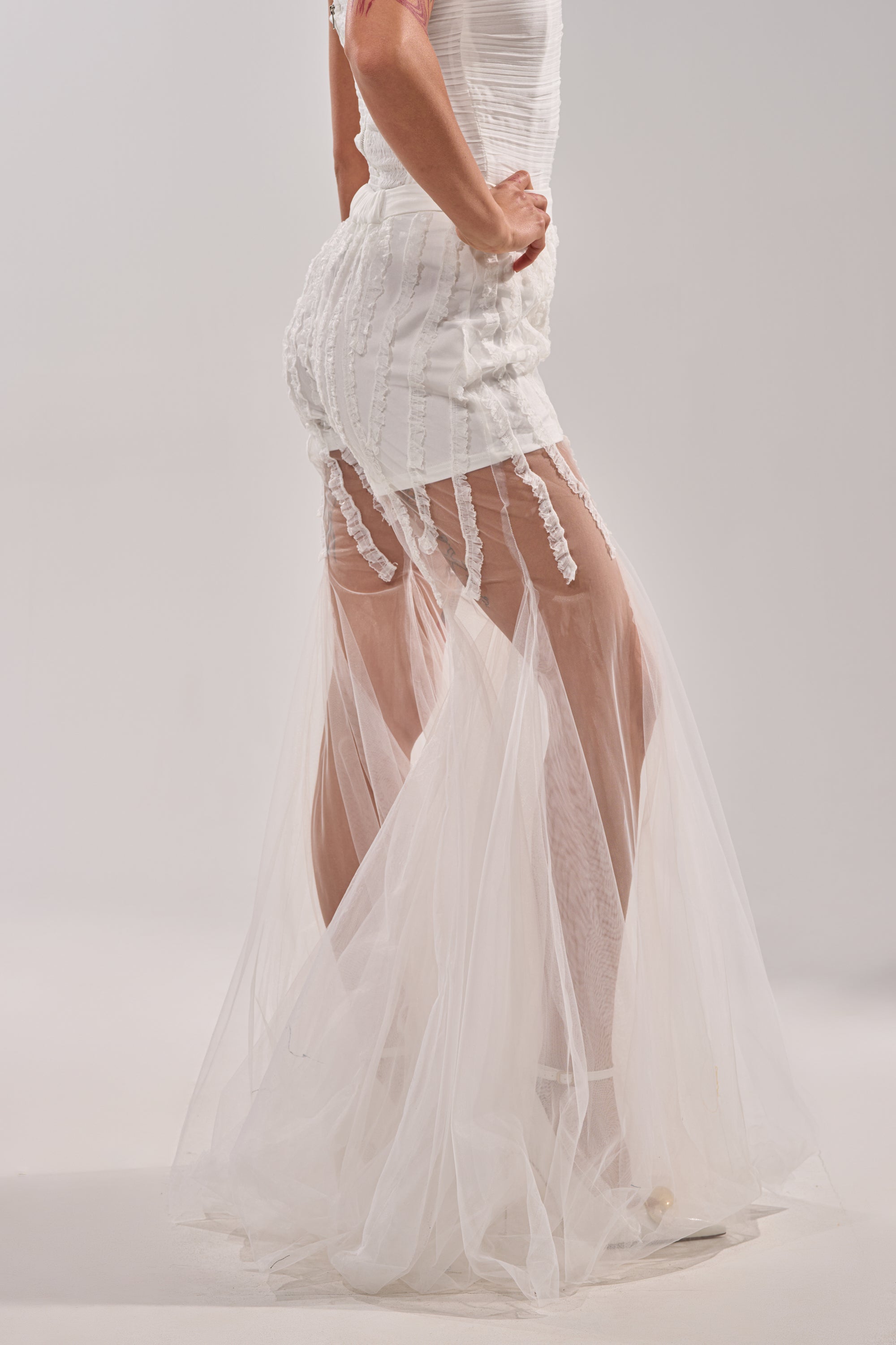 A person wears the EYE FOR MY EYES ON YOUR MESH FLARE PANT, featuring a white fitted look with textured vertical details and a sheer, floor-length tulle overlay. The semi-transparent lower half resembles mesh flare pants against a light background. 2