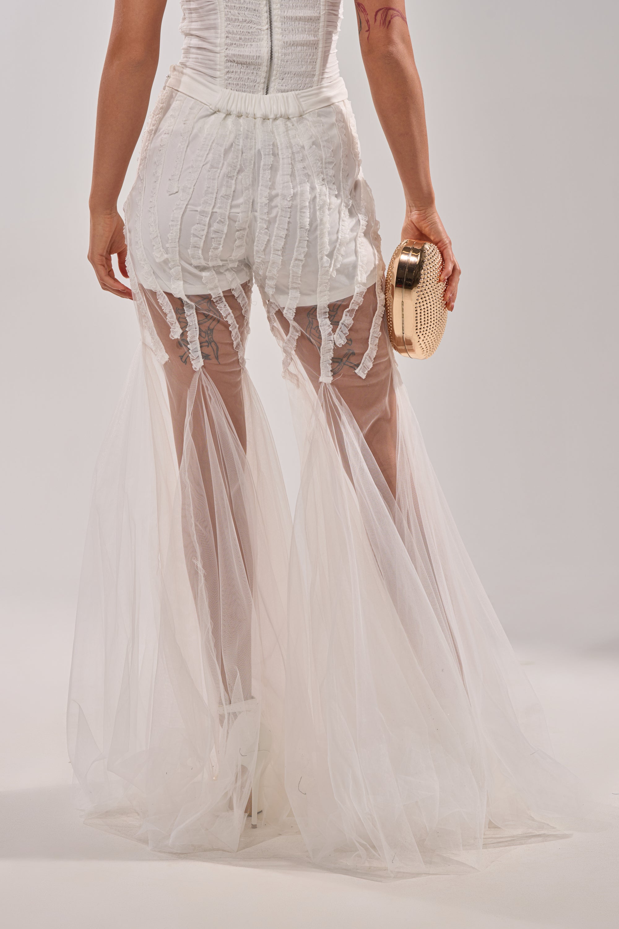 A woman showcases statement style in the EYE FOR MY EYES ON YOUR MESH FLARE PANT with visible shorts, holding a round gold clutch and photographed from the back against a light background. 3