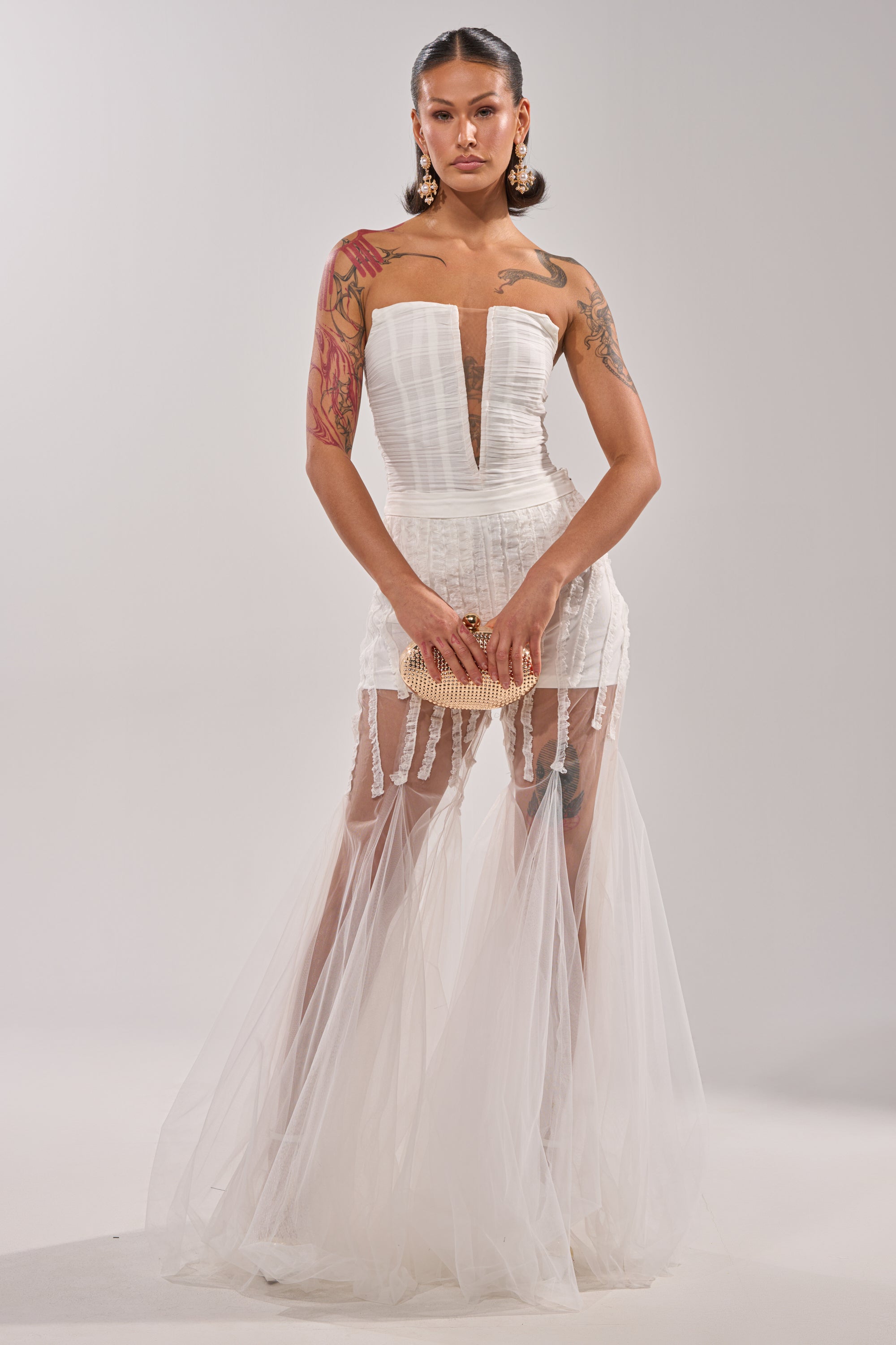 A tattooed woman models the "EYE FOR MY EYES ON YOUR MESH FLARE PANT," pairing it with a strapless white V-neck dress, large gold earrings, and a small textured clutch against a plain light background. 5