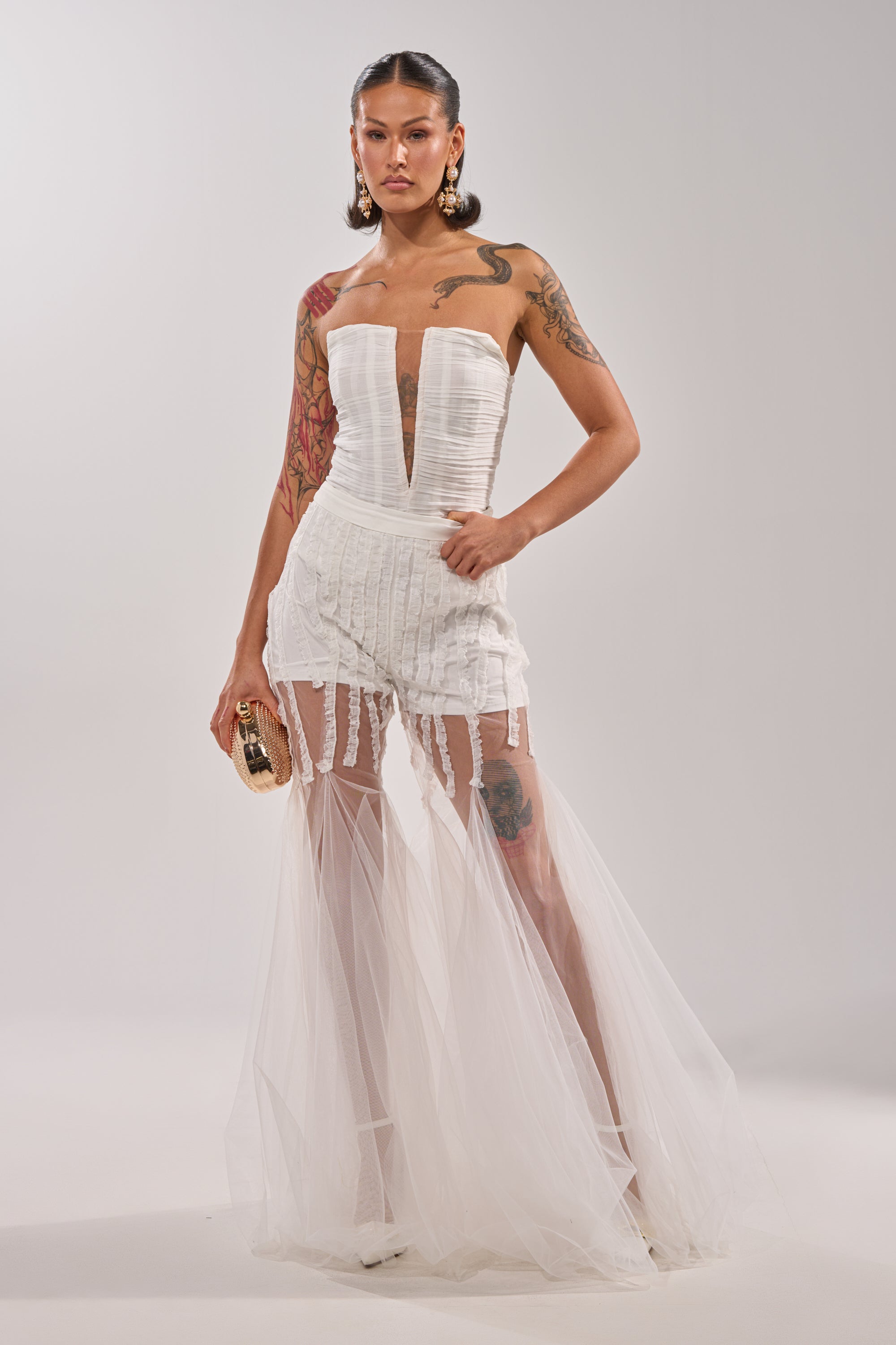 A woman exudes confidence in the EYE FOR MY EYES ON YOUR MESH FLARE PANT, pairing its sheer mesh and bold flare with a gold clutch. Slicked-back hair and visible tattoos complete her daring, statement-making look. 6