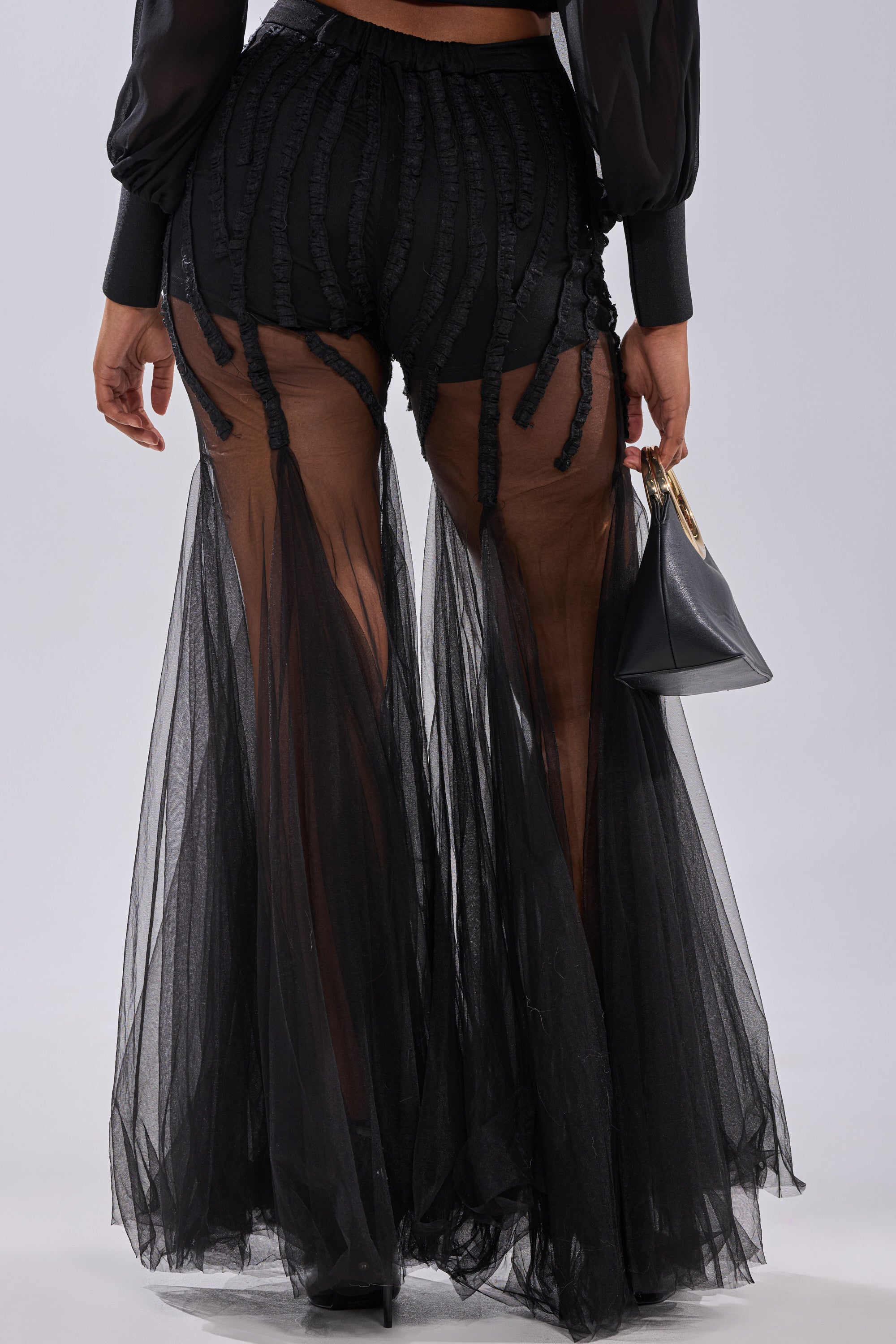 Wearing the KYLIE MESH FLARE PANT—sheer black mesh with ruffle details and built-in shorts—a person holds a small black handbag. The photo is taken from behind, highlighting the lower half of this high-rise flare look.