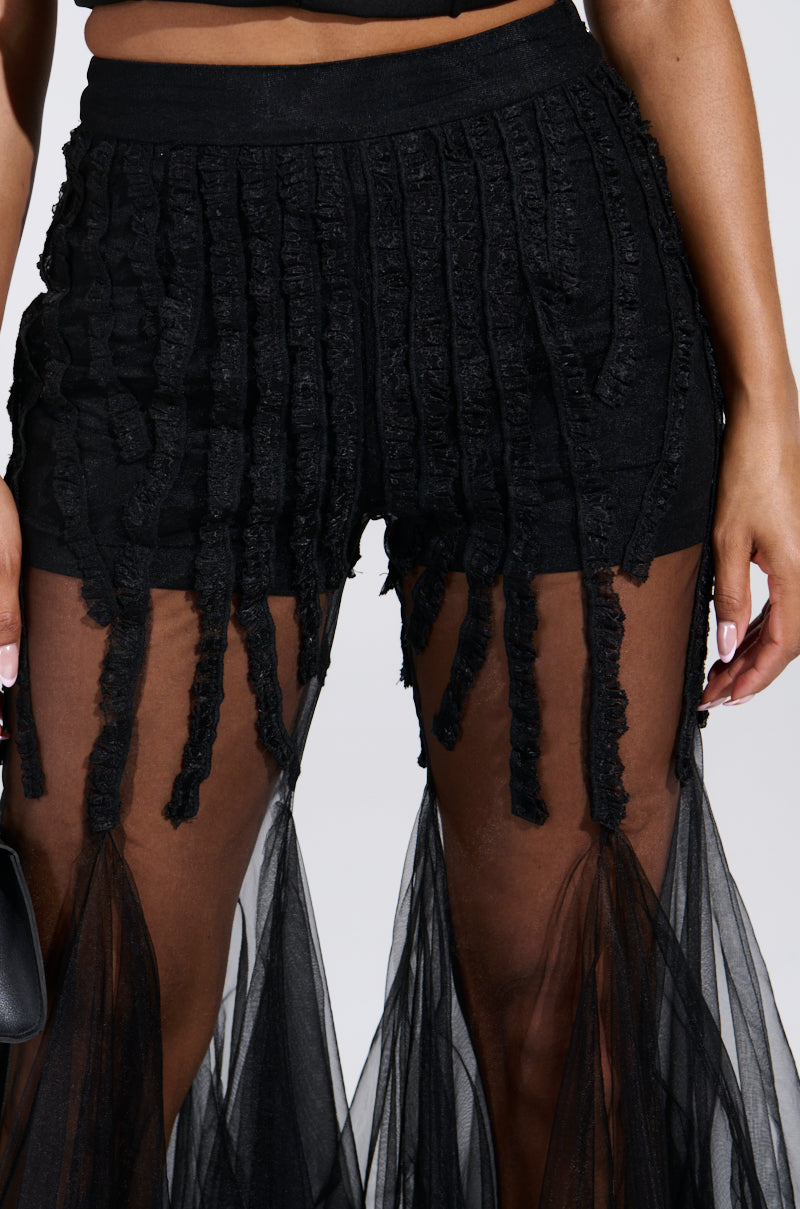 A person is wearing the KYLIE MESH FLARE PANT, featuring textured vertical stripes and sheer mesh flare fabric from the knees for a flowing effect. They have a small black handbag, and their nails are light pink.