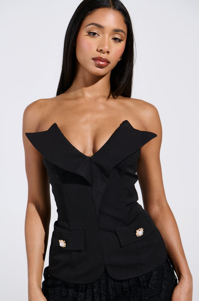 A woman with long hair models the chic KYLIE BLOUSE, featuring an off-shoulder design with a plunging V-neckline, decorative pocket buttons, and a metallic back zipper. The background is plain and well-lit. 0