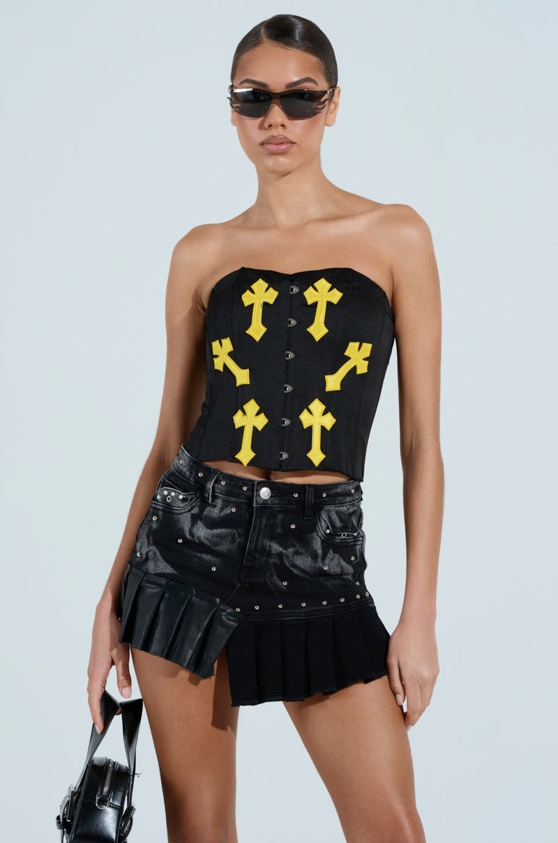 Someone wearing the JAYLIN CROSSED CORSET, a chic strapless black piece with yellow crosses, paired with a black pleated mini skirt and dark sunglasses. They hold a sleek black handbag, all set against a plain white background.