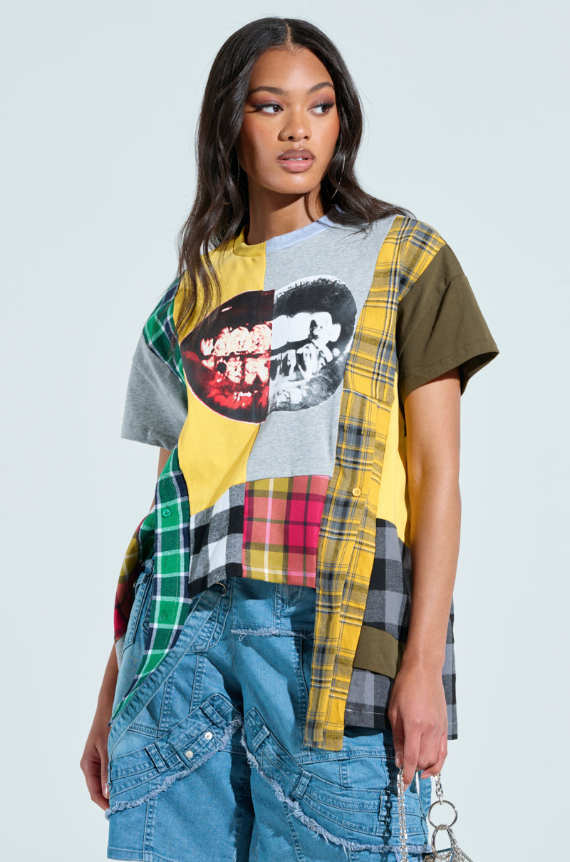 A person wears a REBUILT OVERSIZED FLANNEL T-SHIRT with a patchwork design and smiling mouth graphic. Long hair flows as they hold a chain. Their jeans have decorative stitching, set against a plain, light-colored background.