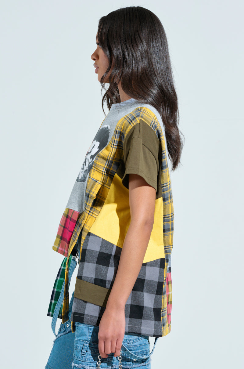 With long dark hair, a person stands in profile wearing the REBUILT OVERSIZED FLANNEL T-SHIRT, showcasing a patchwork design with plaid and solid patterns in shades of yellow, red, black, and gray. They pair it with light blue jeans while holding sunglasses in their right hand.