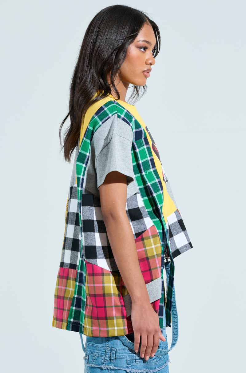 A person in a REBUILT OVERSIZED FLANNEL T-SHIRT with multicolored, checkered vest details and patchwork design over gray and blue jeans stands against a light background. They have long, dark hair.