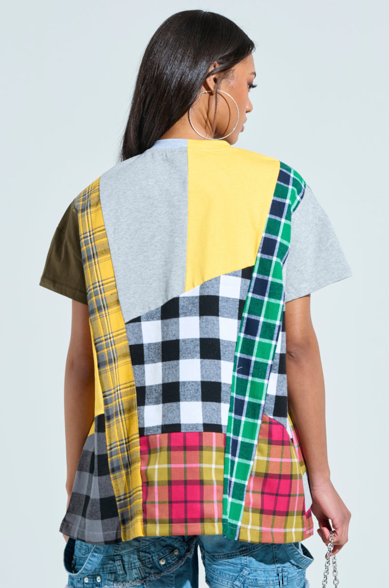 A person with long hair is wearing an oversized REBUILT FLANNEL T-SHIRT featuring a vibrant plaid and check patchwork design in gray, yellow, green, black-and-white, and red on the back, complemented by blue jeans.