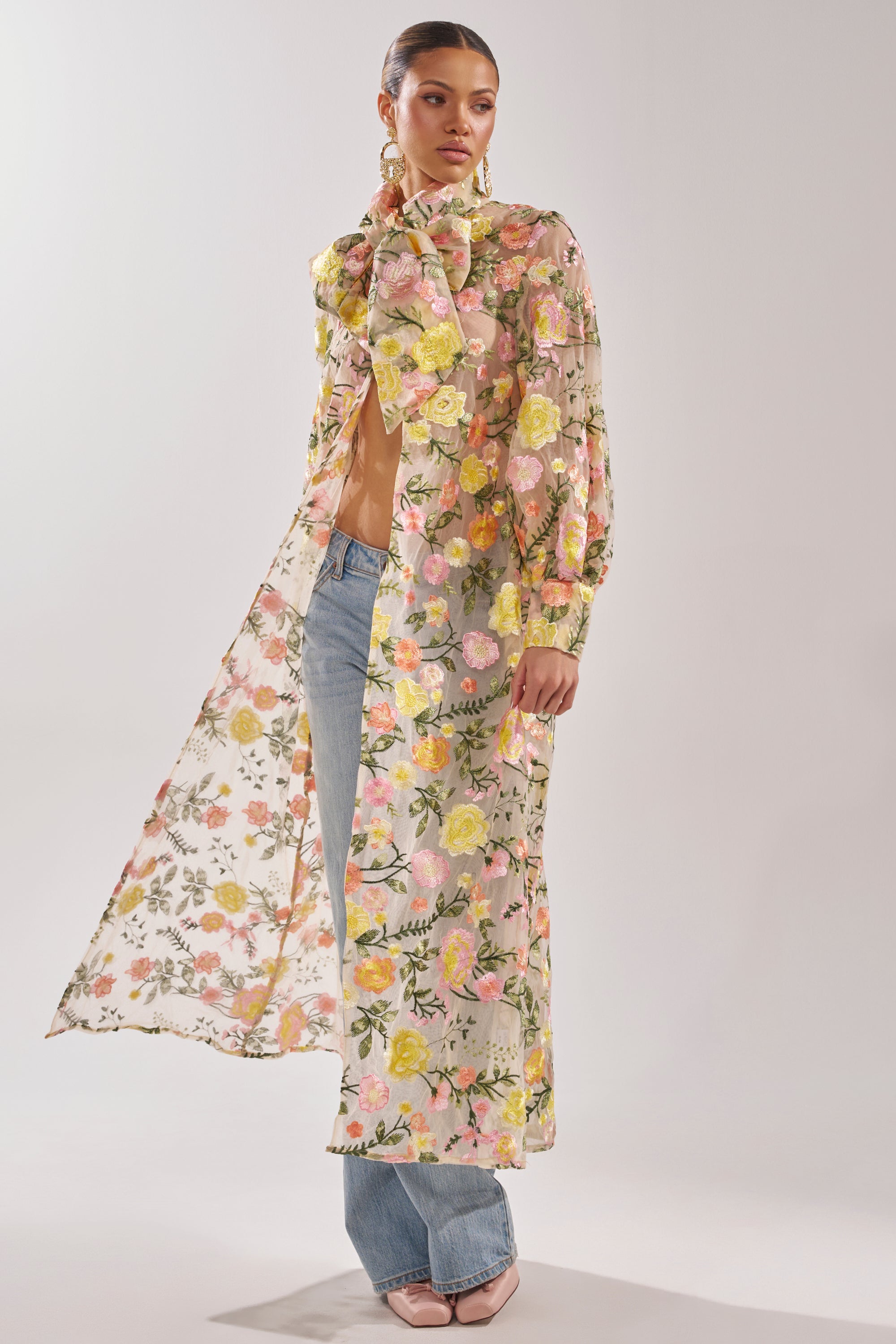 A woman poses against a plain background wearing the BOUQUET DREAMS FLORAL TULLE DUSTER with a large neck bow, paired with blue jeans and nude heels.