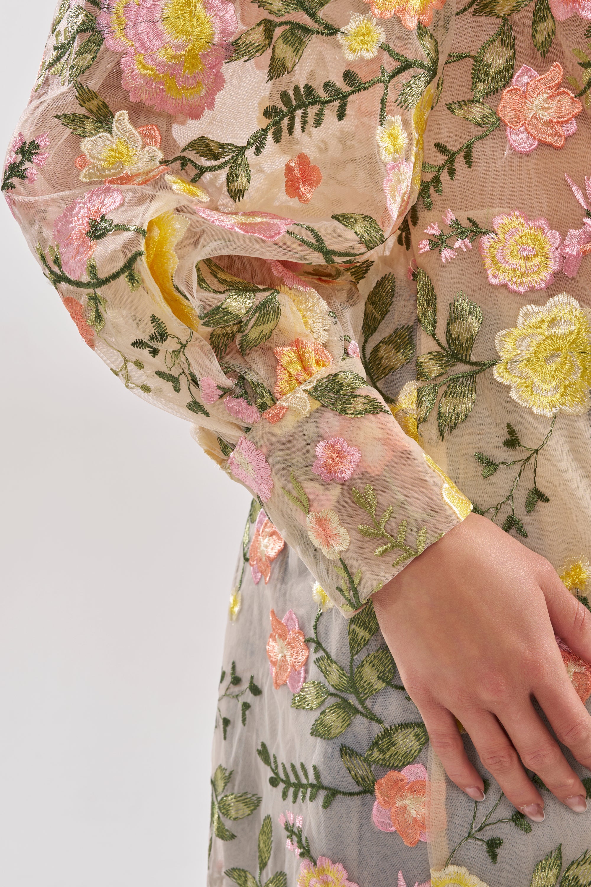 A close-up of a hand resting on the side, wearing the BOUQUET DREAMS FLORAL TULLE DUSTER—a sheer maxi duster with colorful pink, yellow, and green floral embroidery on light fabric.
