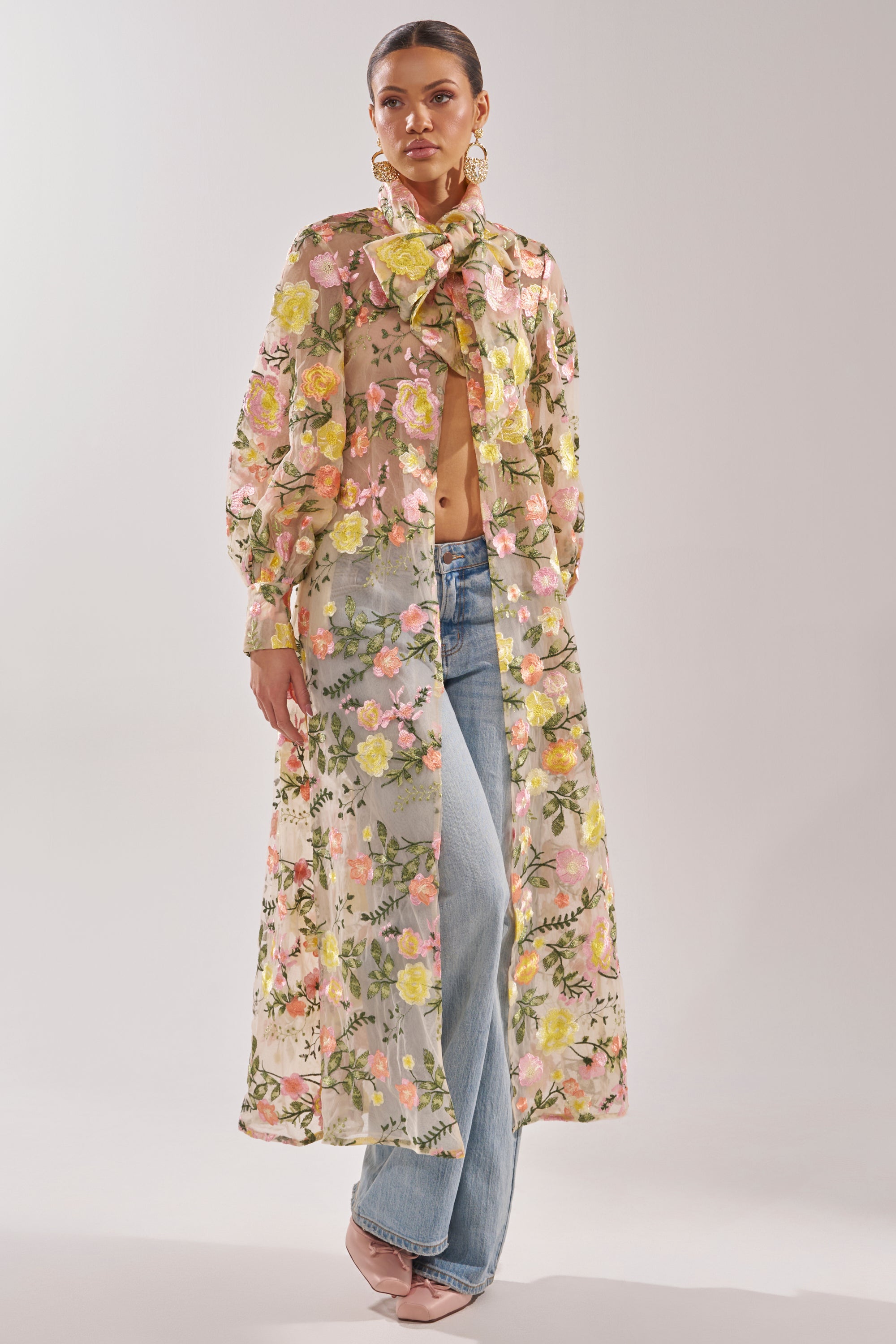 A woman wears the BOUQUET DREAMS FLORAL TULLE DUSTER—a sheer maxi coat with pastel pink, yellow, and green embroidered flowers and a statement neck bow—styled with light blue jeans and pale heels against a plain, light background.