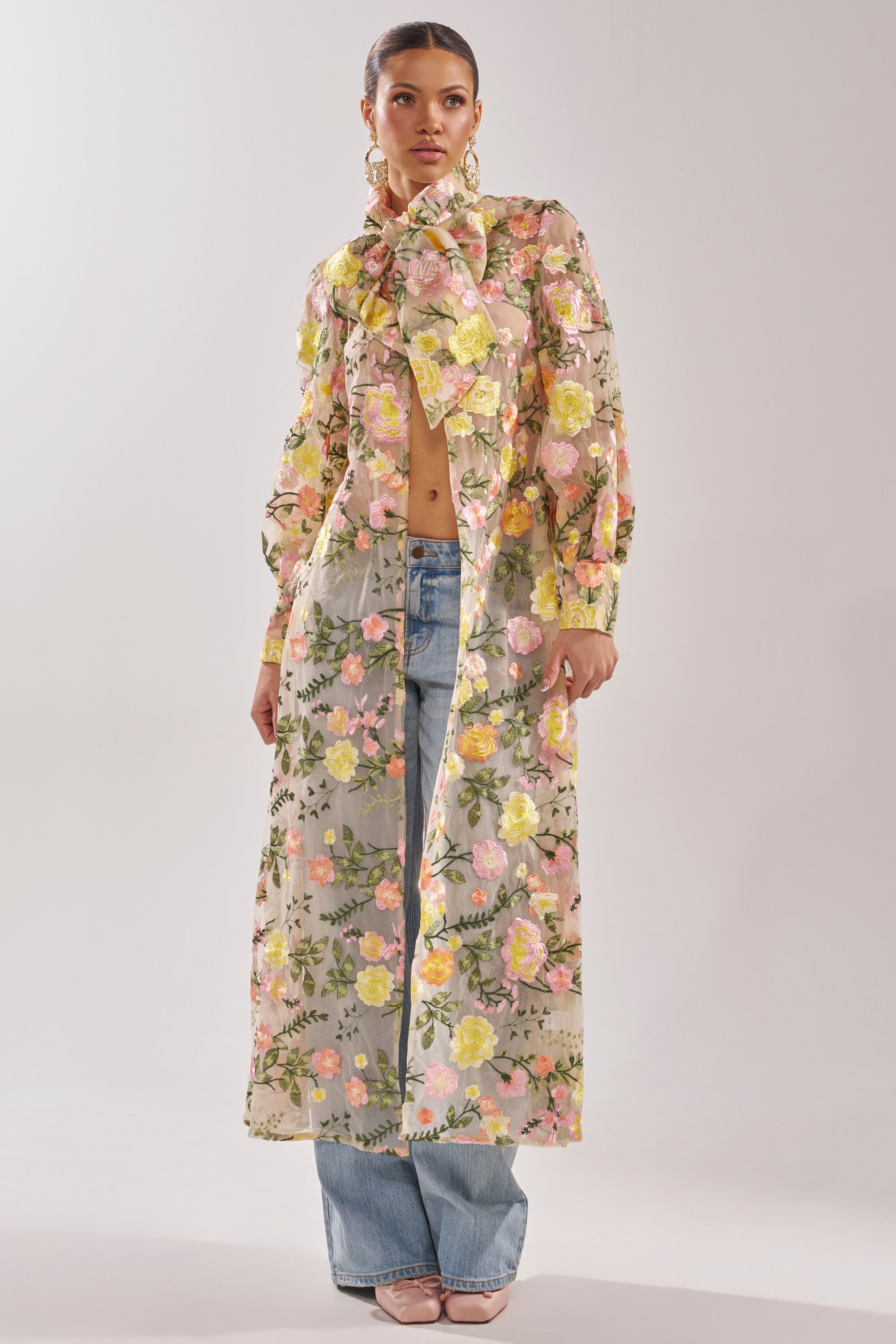A woman wears the BOUQUET DREAMS FLORAL TULLE DUSTER—a sheer maxi coat with a large neck bow and pastel floral embroidery—styled over light blue jeans and pale pink heels, revealing part of her torso through the translucent fabric.